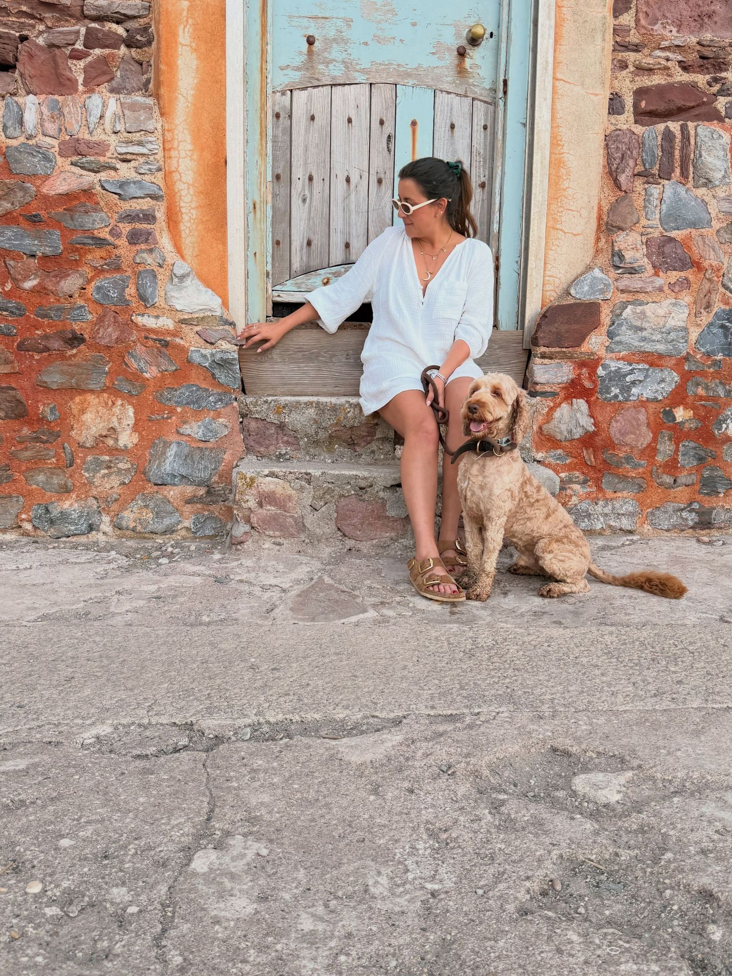 Woman in white dress sitting on stone steps with a fluffy golden doodle dog beside a rustic turquoise door.