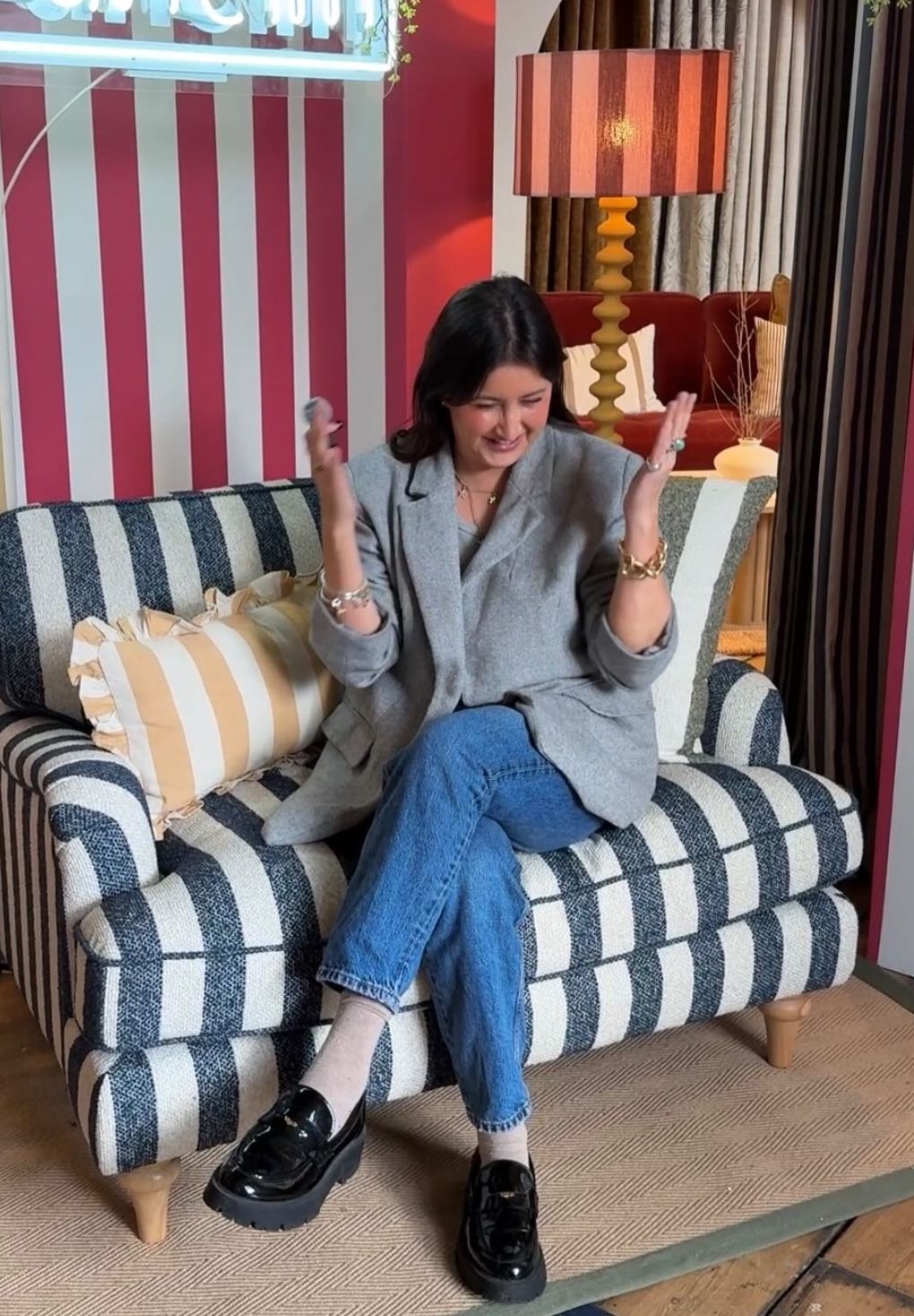 Woman in gray blazer and jeans sits on striped sofa, smiling with hands raised in a colorful interior.