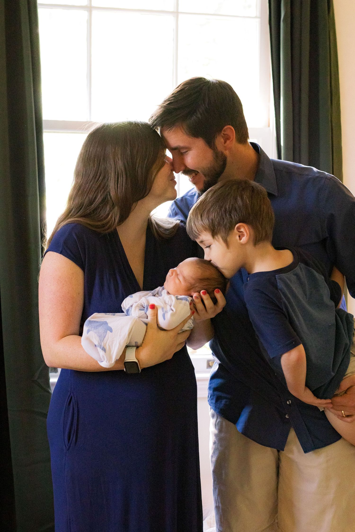 A beautiful family of four in from of a window. Mom and dad are kissing. The dad is holding a young boy who is kissing the newborn son.