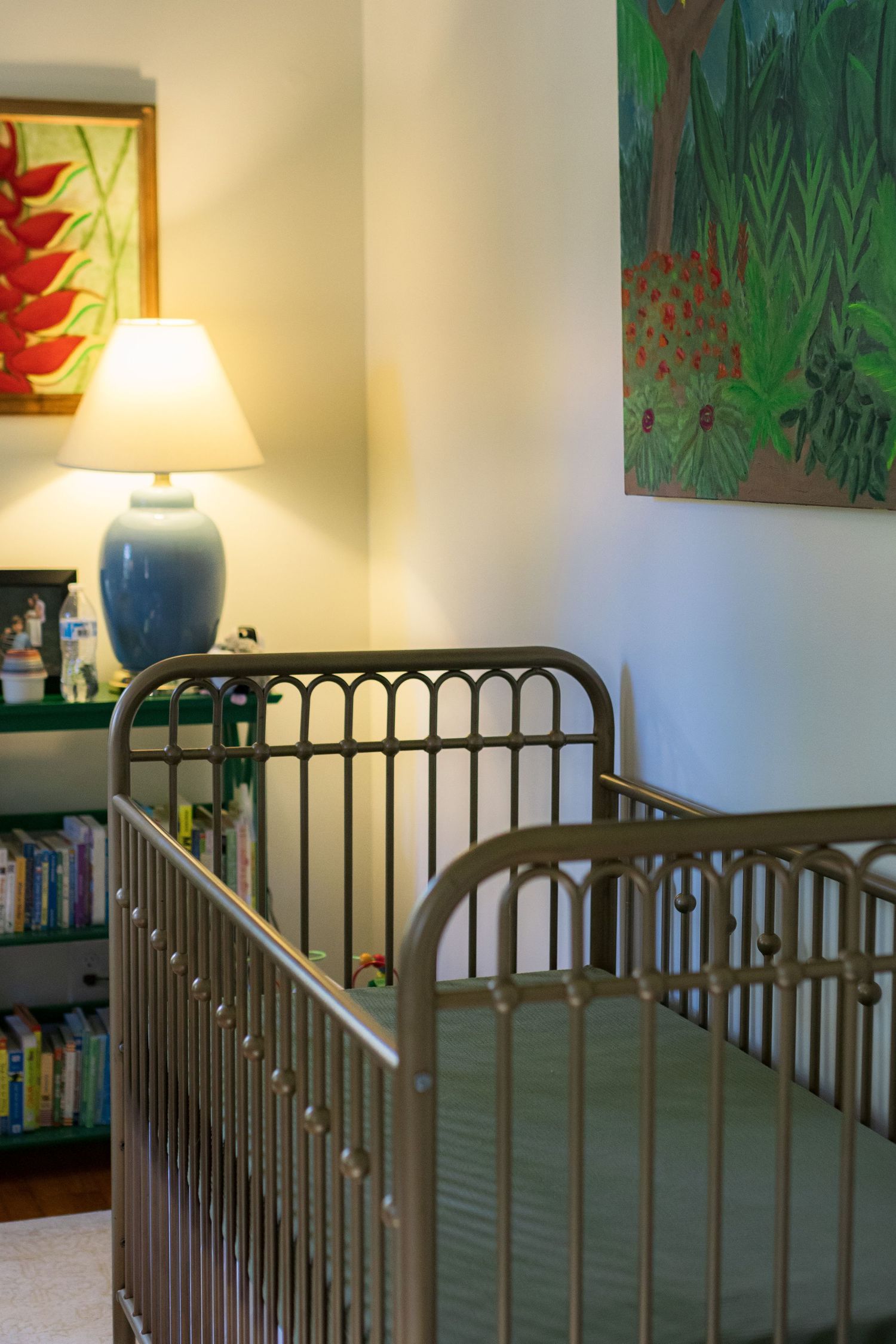 A nursery room with a gold crib with green sheet, paintings on the wall and a blue lamp with a yellow light.