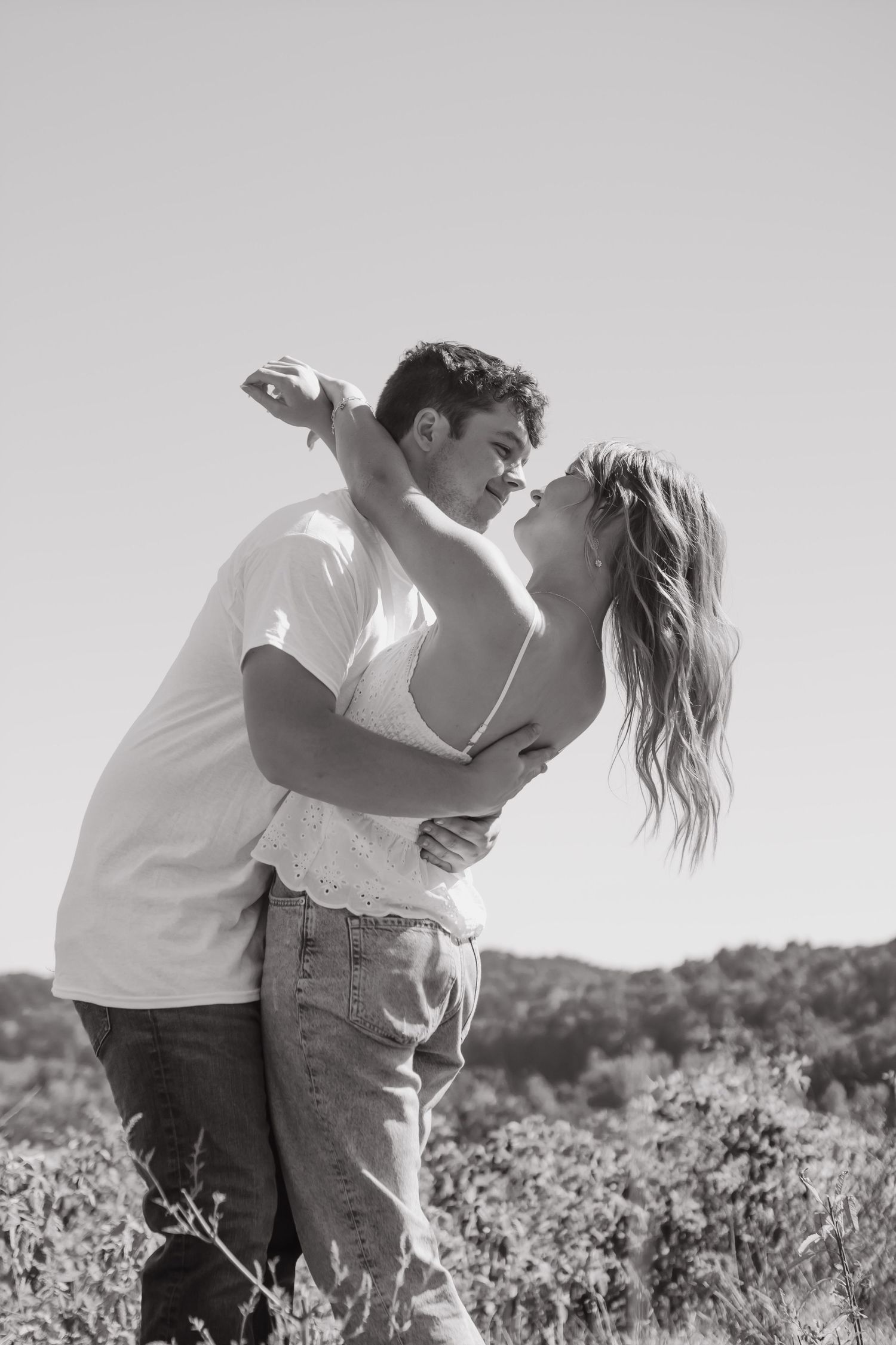 An engaged couple hugging in a field with mountain view and beautiful blue sky. The woman's arms are wrapped around the man's neck.