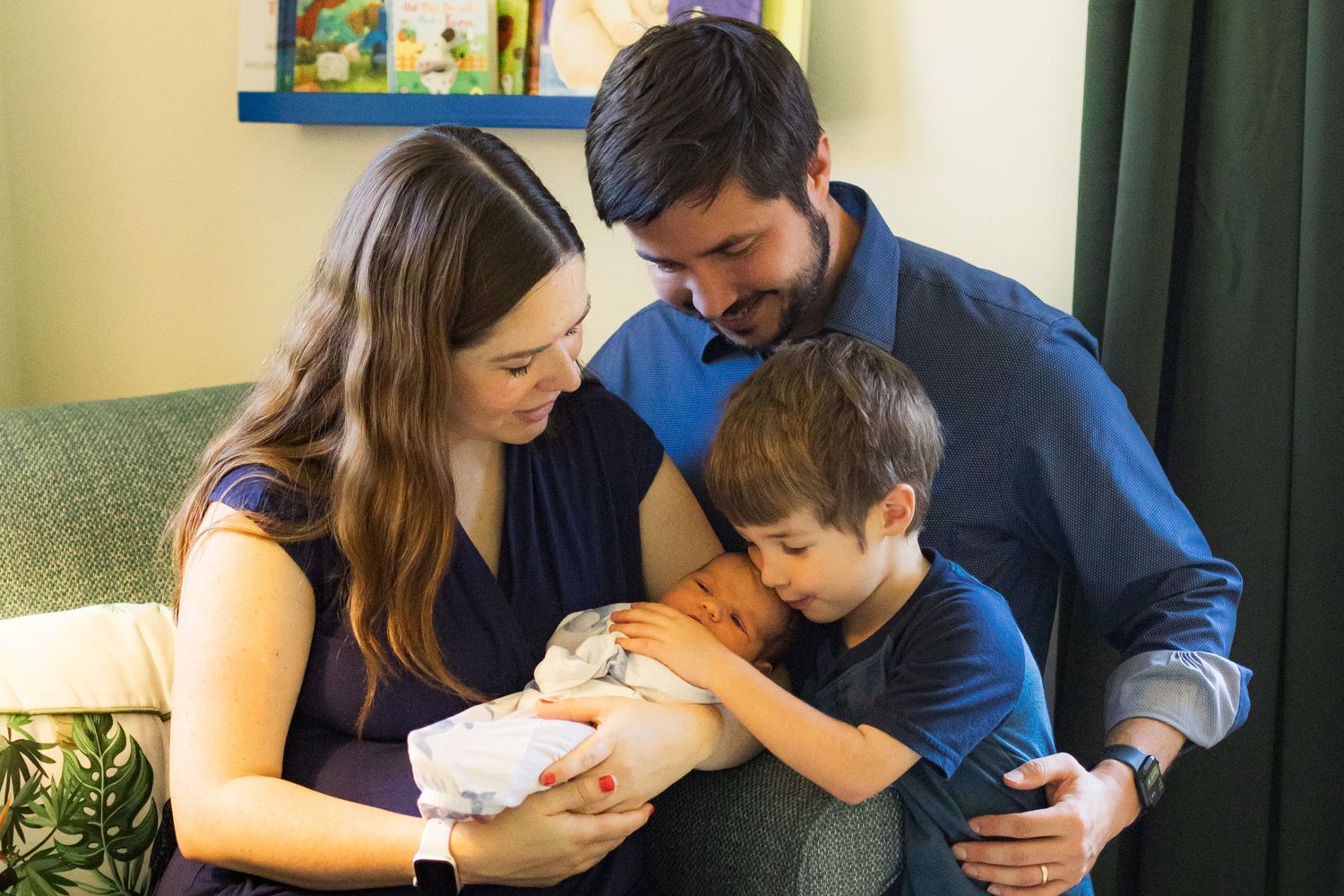 A lovely family of four including gathered together in a nursery. A mom, dad, and four year old son are all looking at a newborn baby.