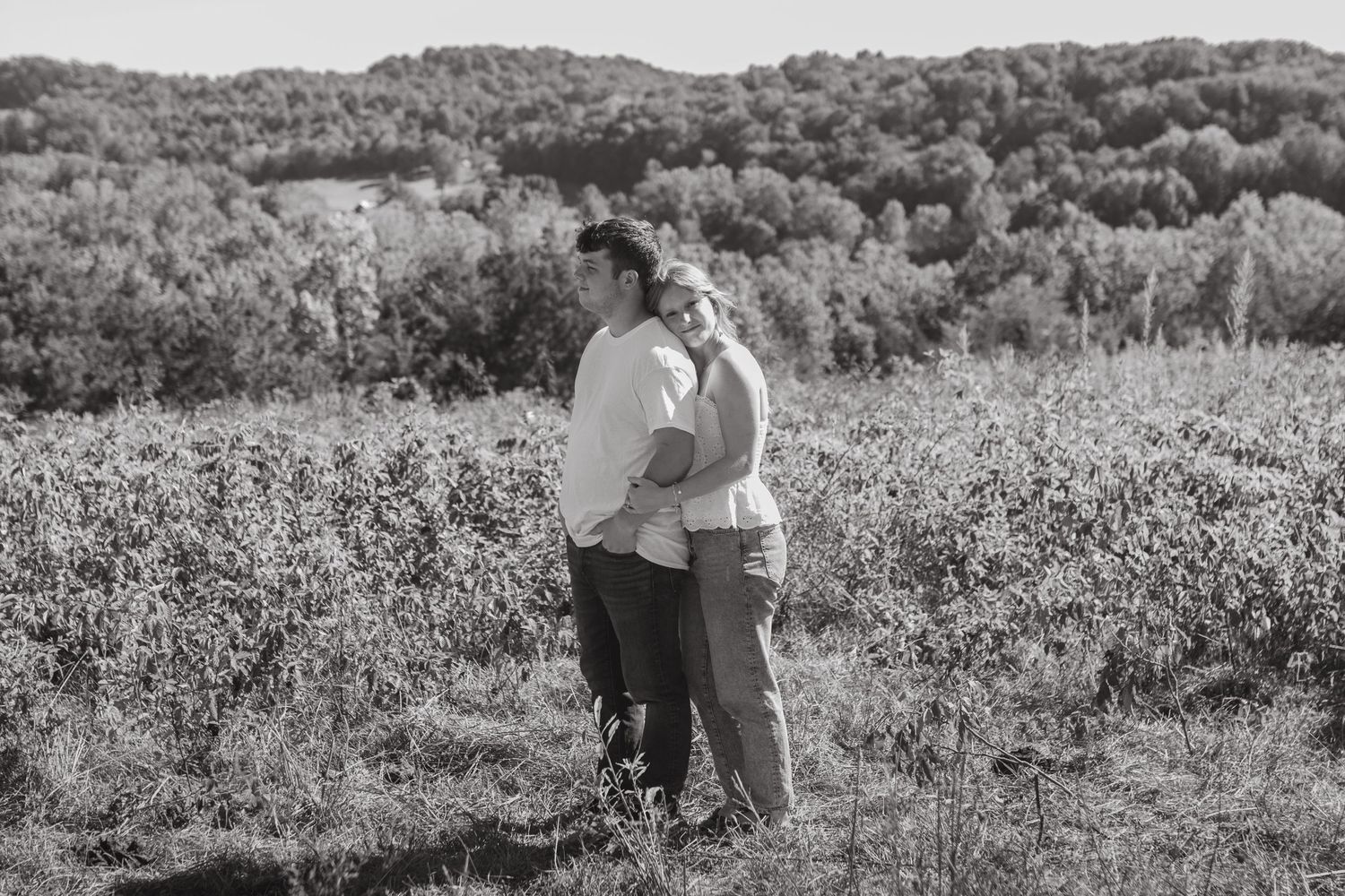 An engaged couple standing in a field with mountain view and beautiful blue sky. The girl hugging the boy and smiling at the camera.