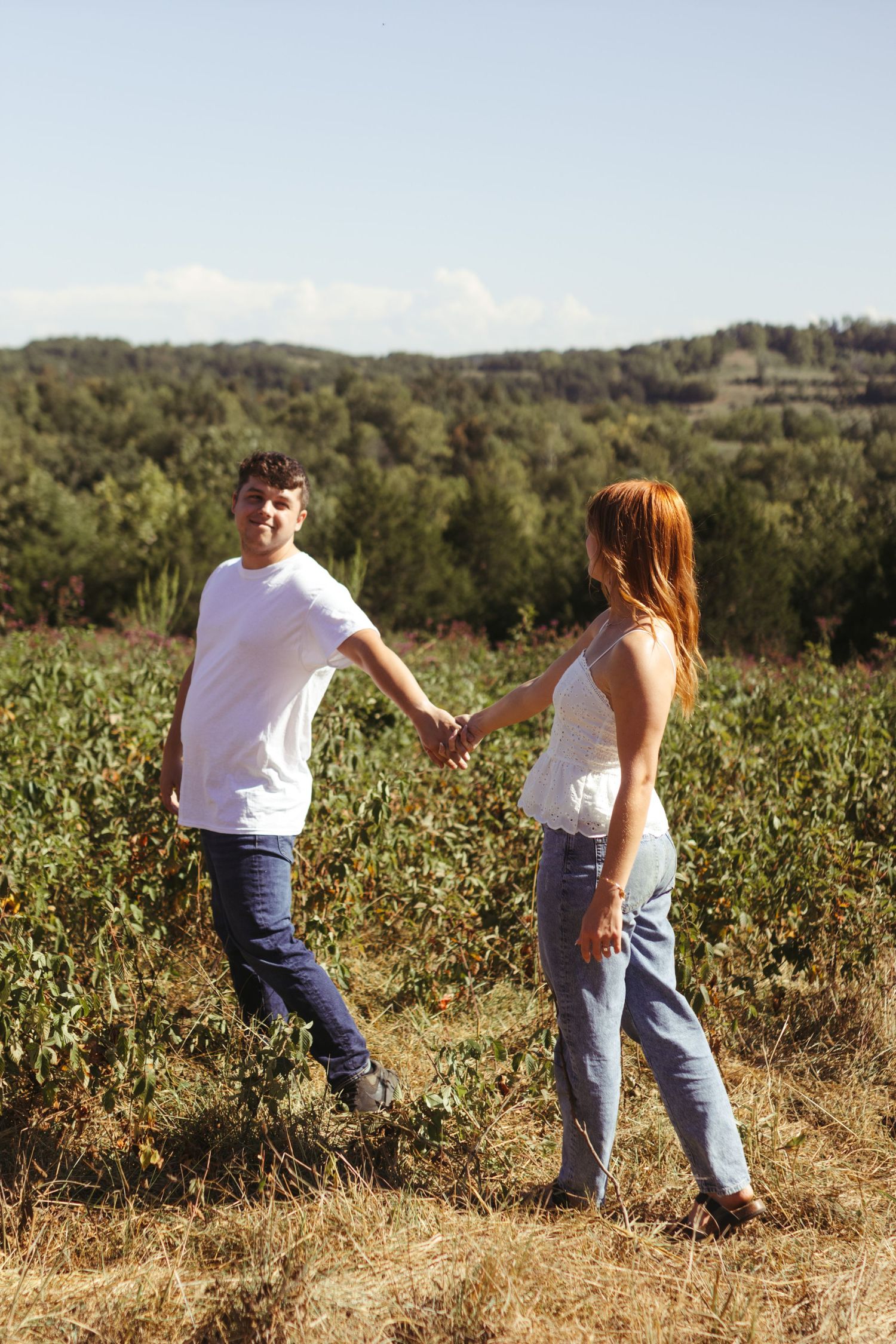 An engaged couple walking in a field with mountain view and beautiful blue sky. The couple is holding hands.