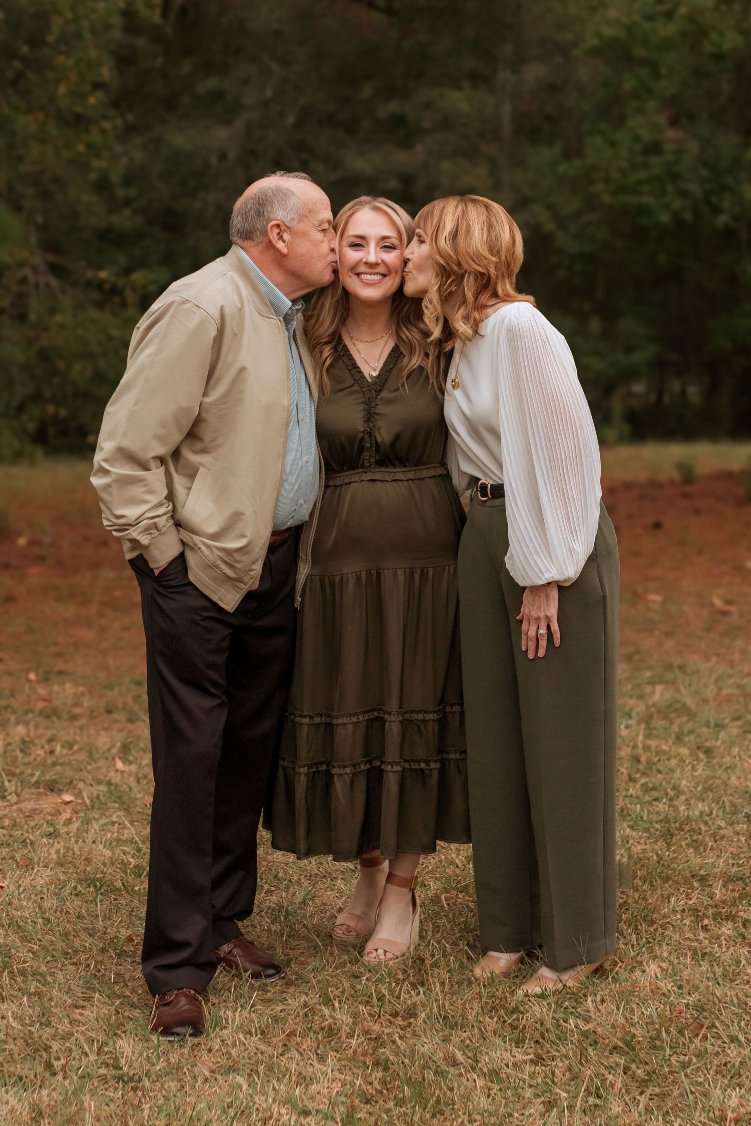 Parents in a field on either side of their daughter leaning in to kiss her on the cheek, arms wrapped around her back.