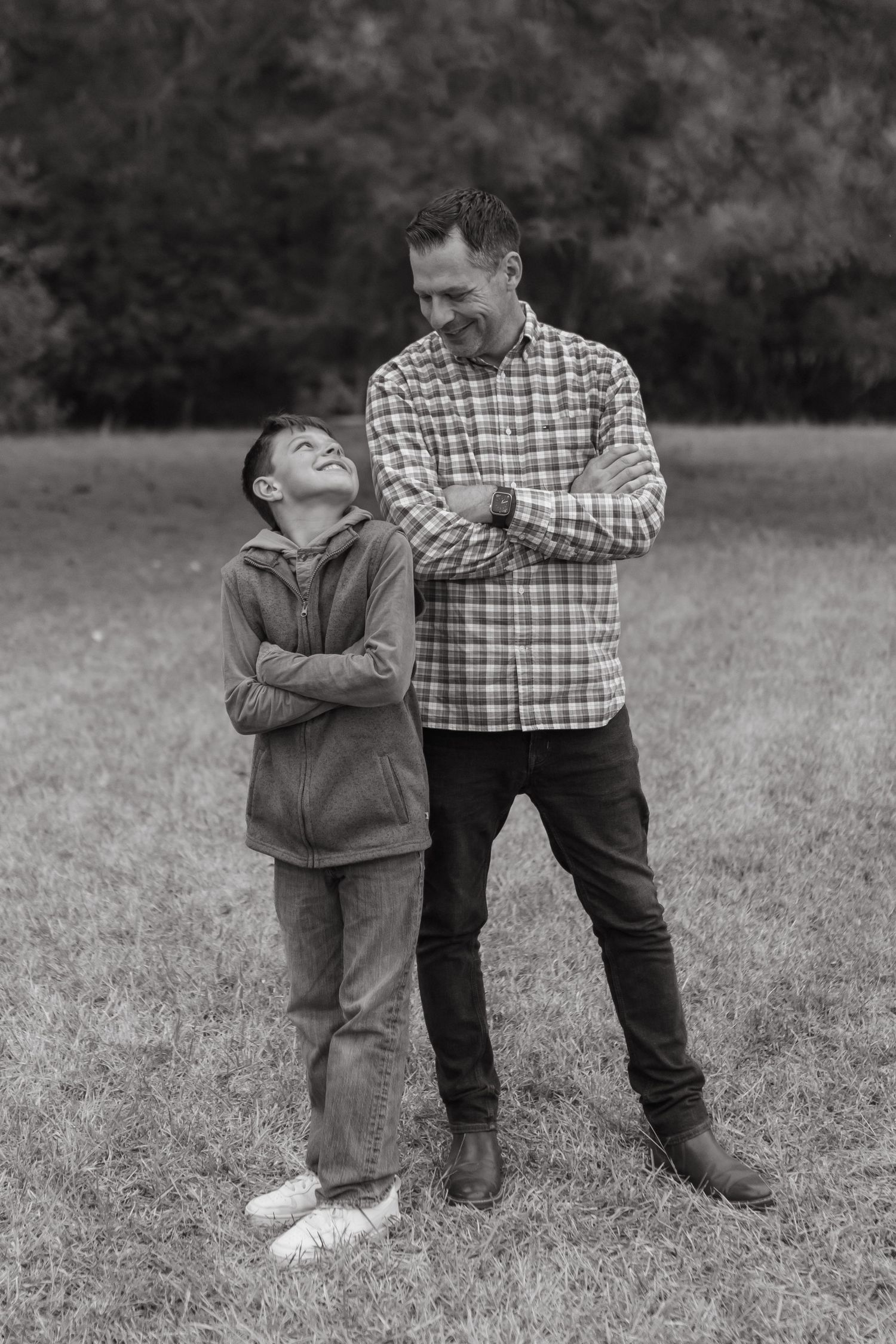 A dad and his son in a field with their arms crossed smiling and looking at each other.