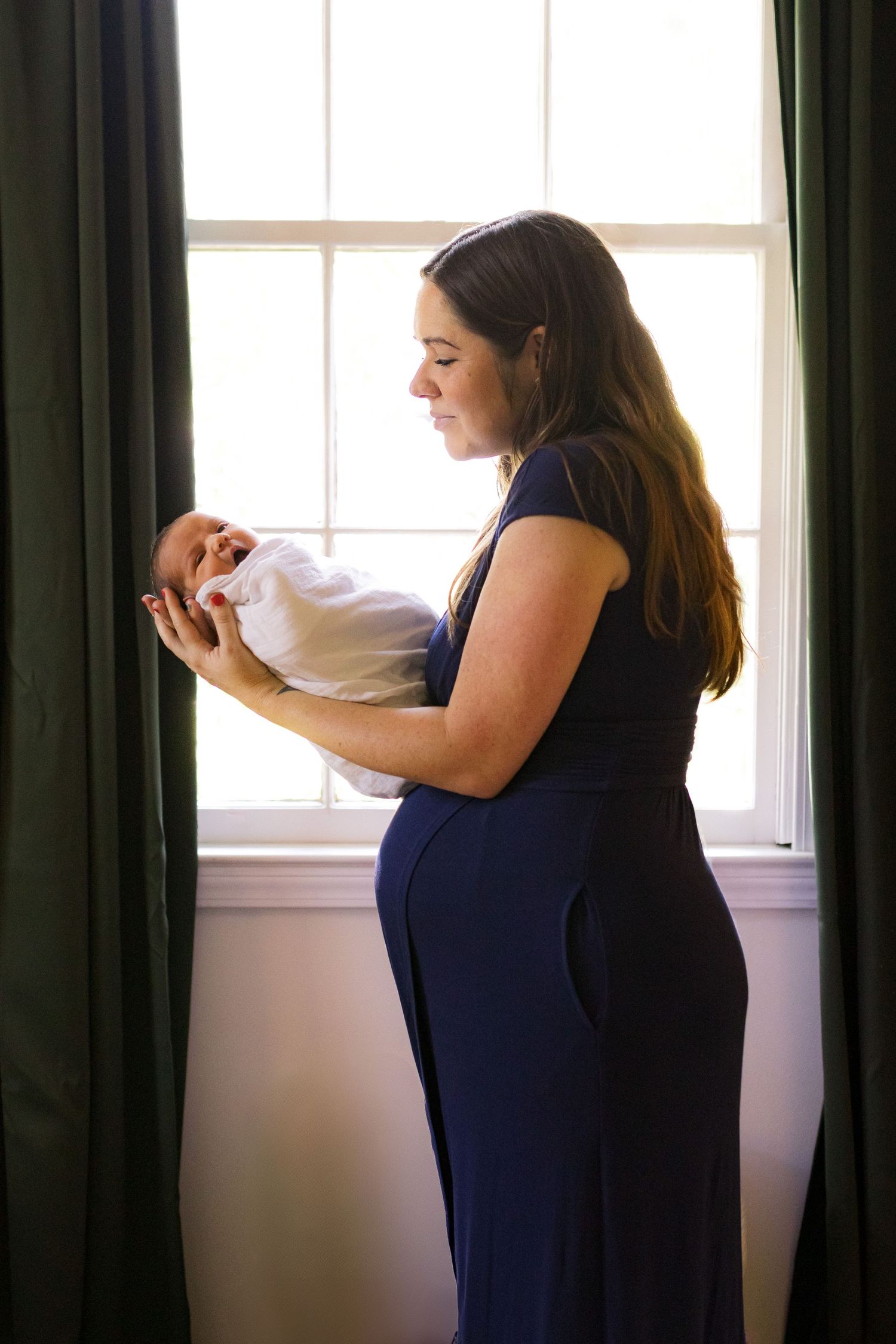 A mom in a nursery in front of a window holding her newborn son and looking at him.
