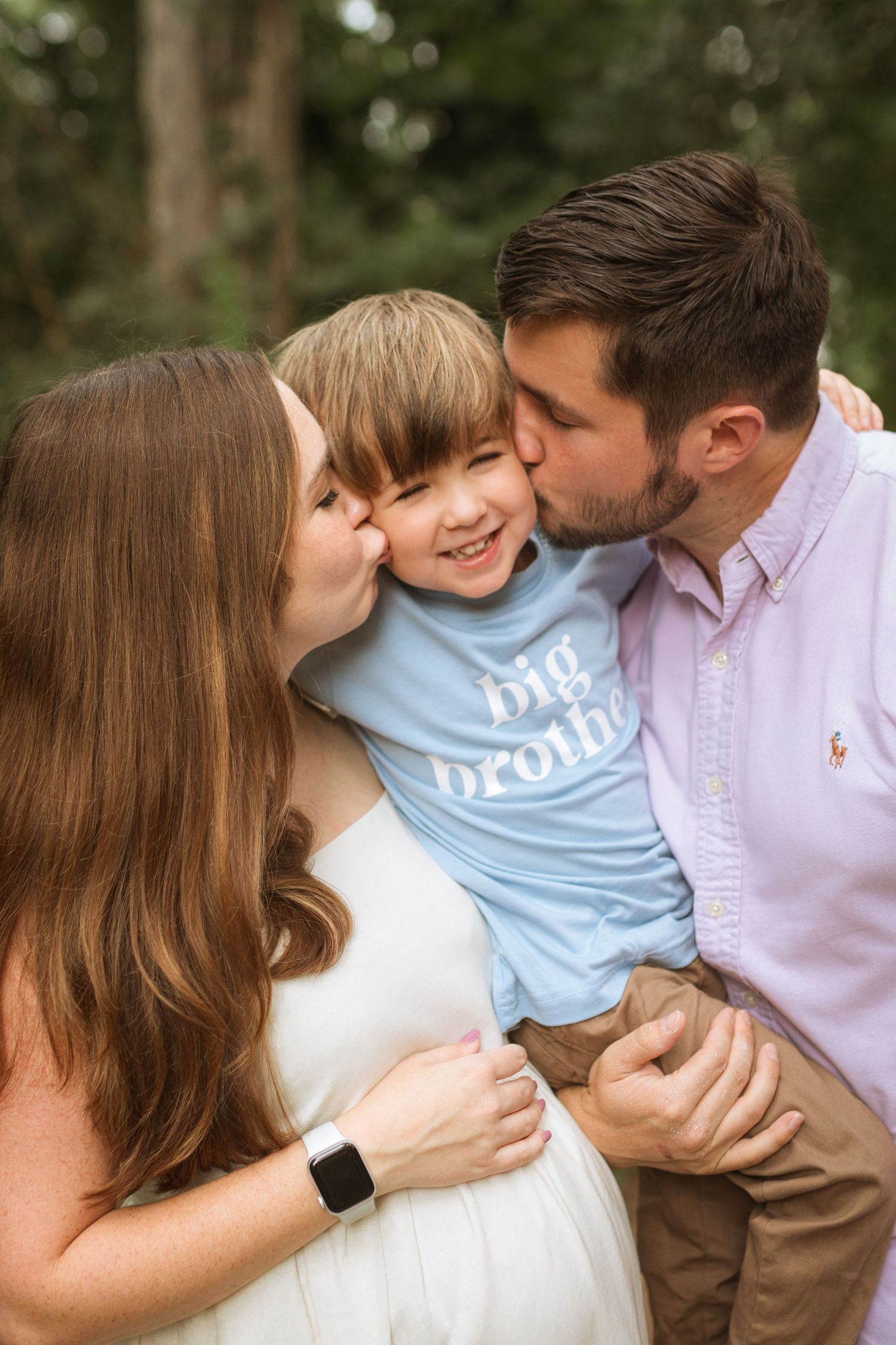 A mom and dad leaning in to kiss their smiling four year old son who has a shirt that reads 