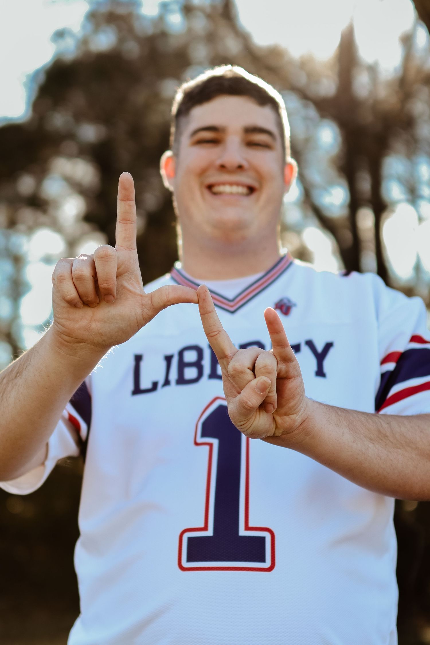 College senior portrait in the woods. Smiling at the camera and making a Liberty University hand symbol.