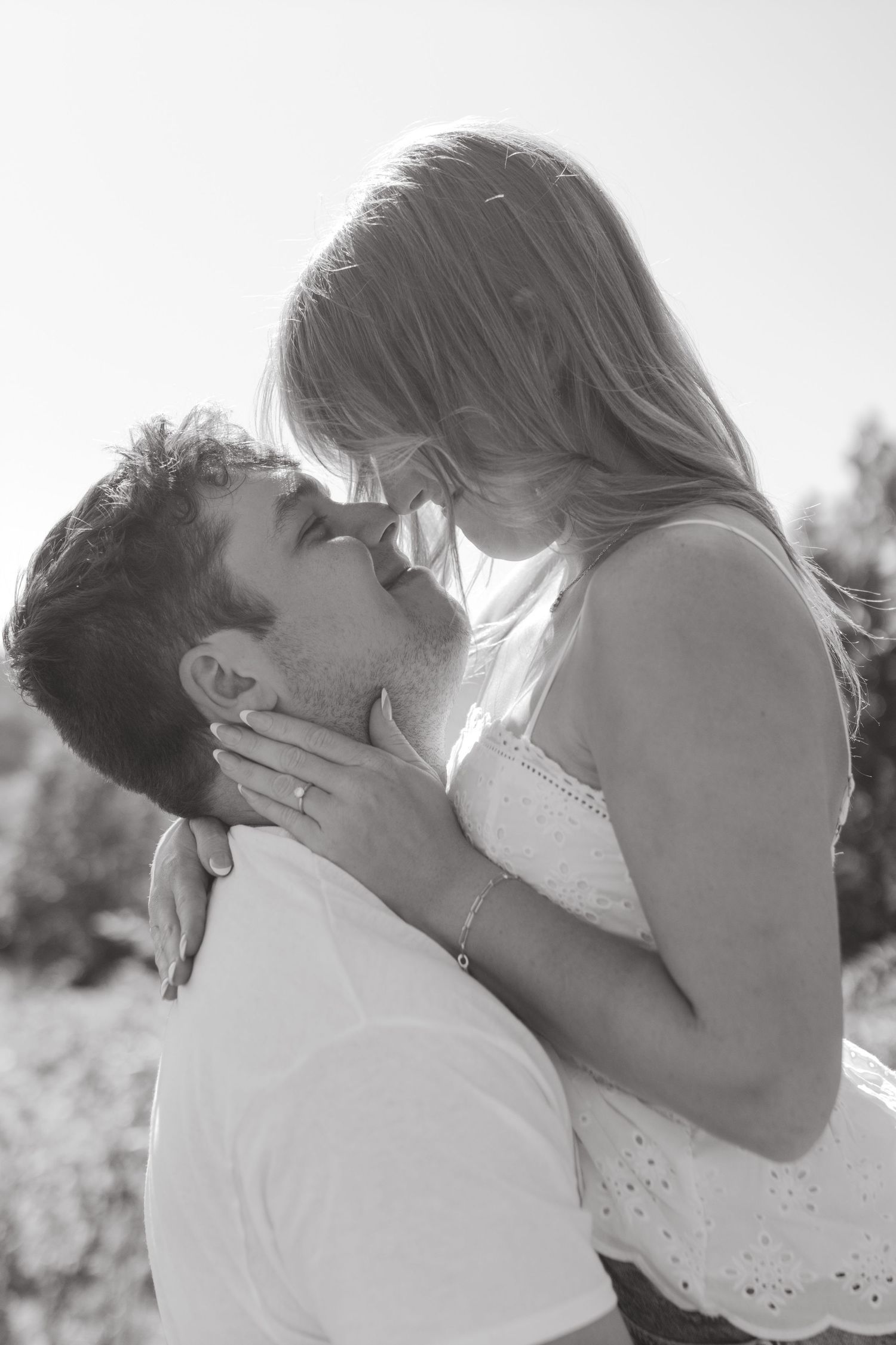 A photo of an engaged couple in a field. The man is picking up the woman and the woman is looking down at the man with her hand on his neck.
