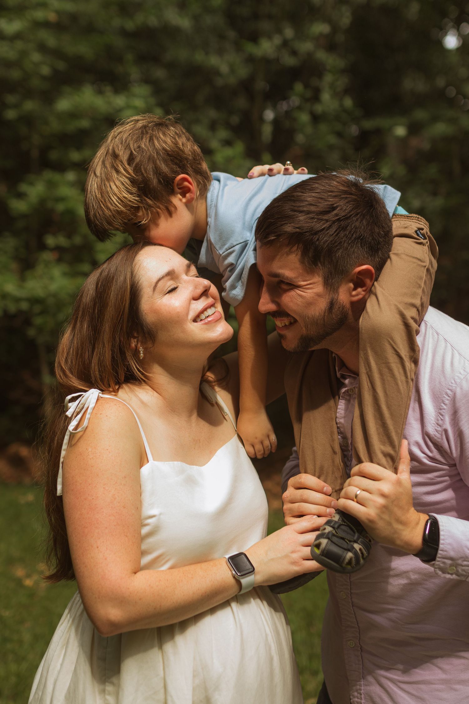 A family photo with a young son on dad's shoulders leaning over to kiss mom on the forehead. The mom is pregnant.