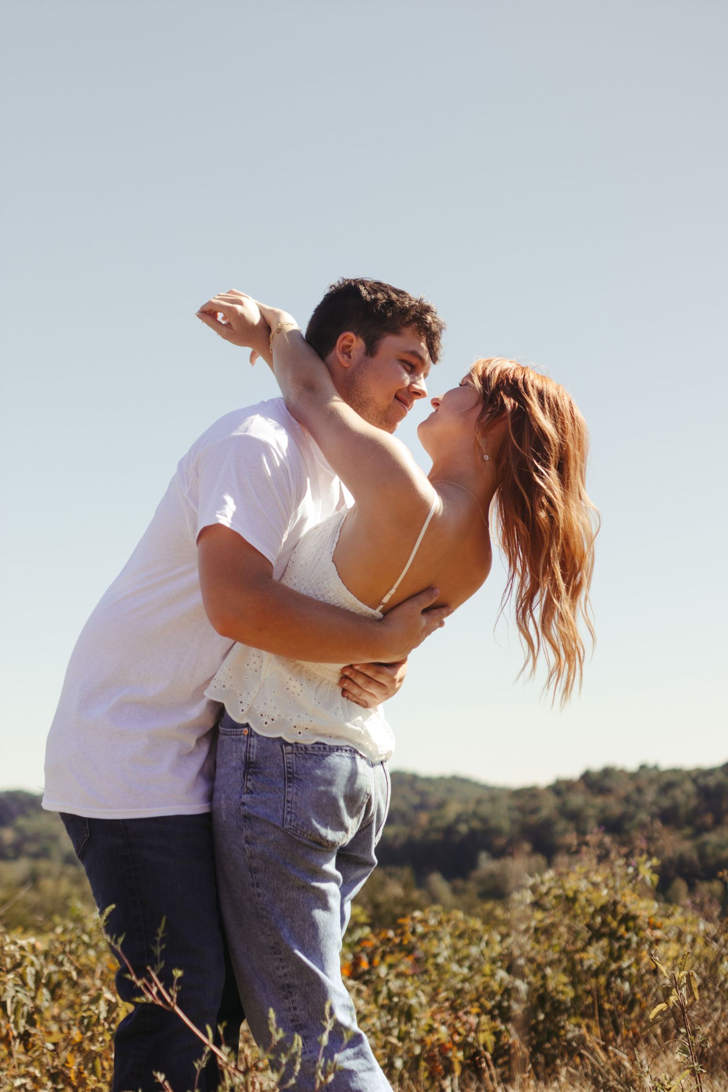 An engaged couple hugging in a field with mountain view and beautiful blue sky. The woman's arms are wrapped around the man's neck.