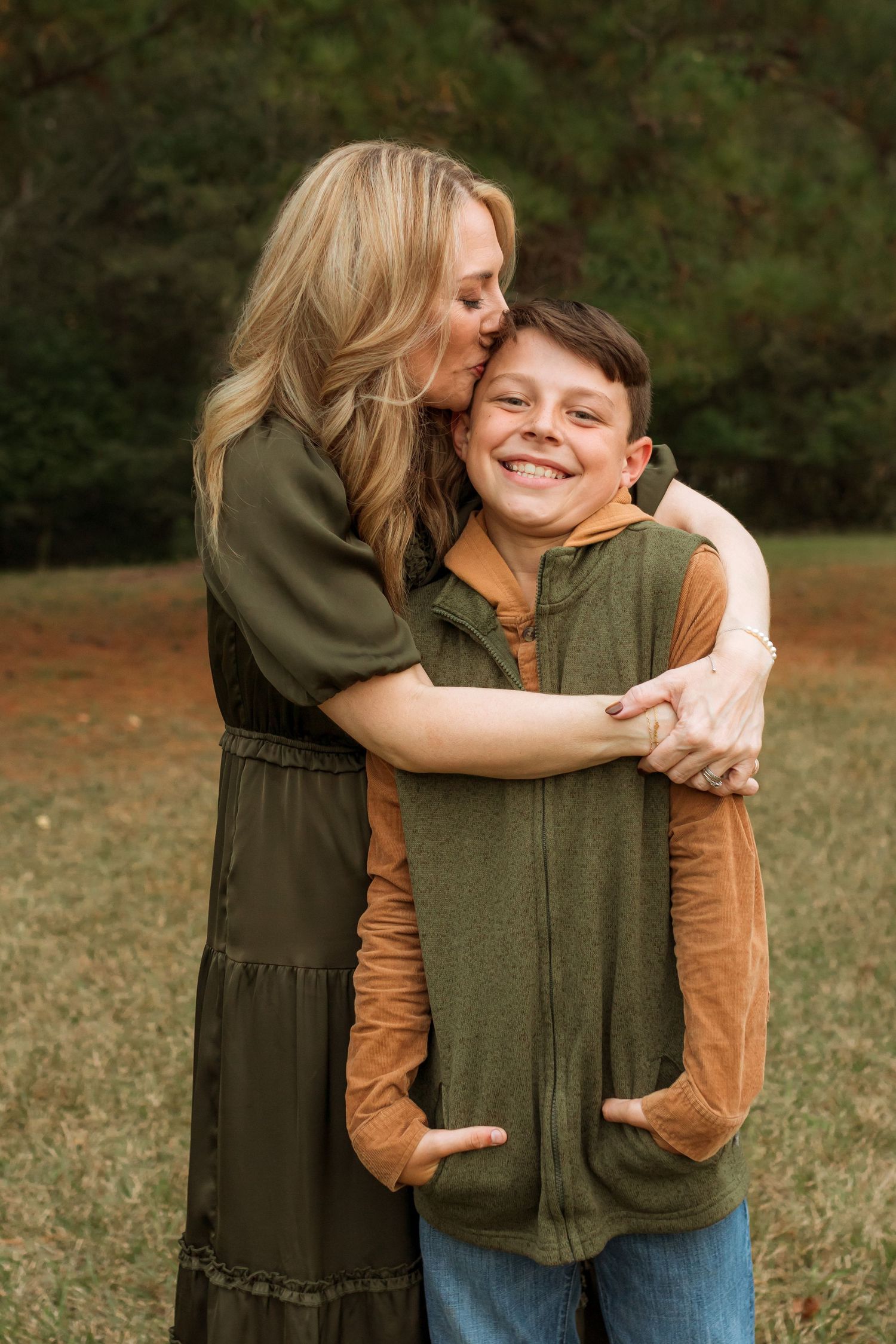 A mom in a field hugging and kissing her 10 year old son on the forehead.