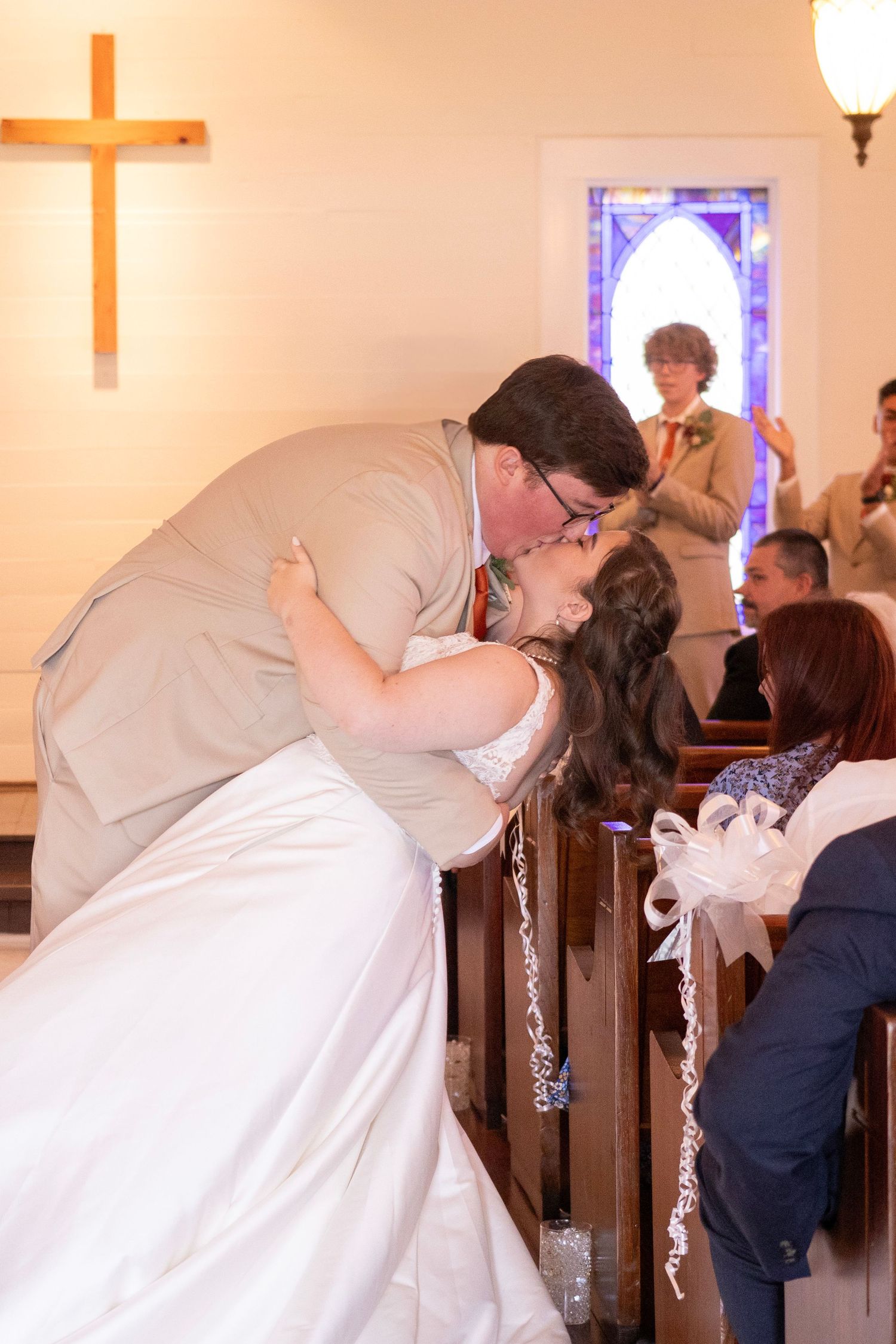 Wedding day kiss at the end of the ceremony. Cross hung up on the wall, people cheering.