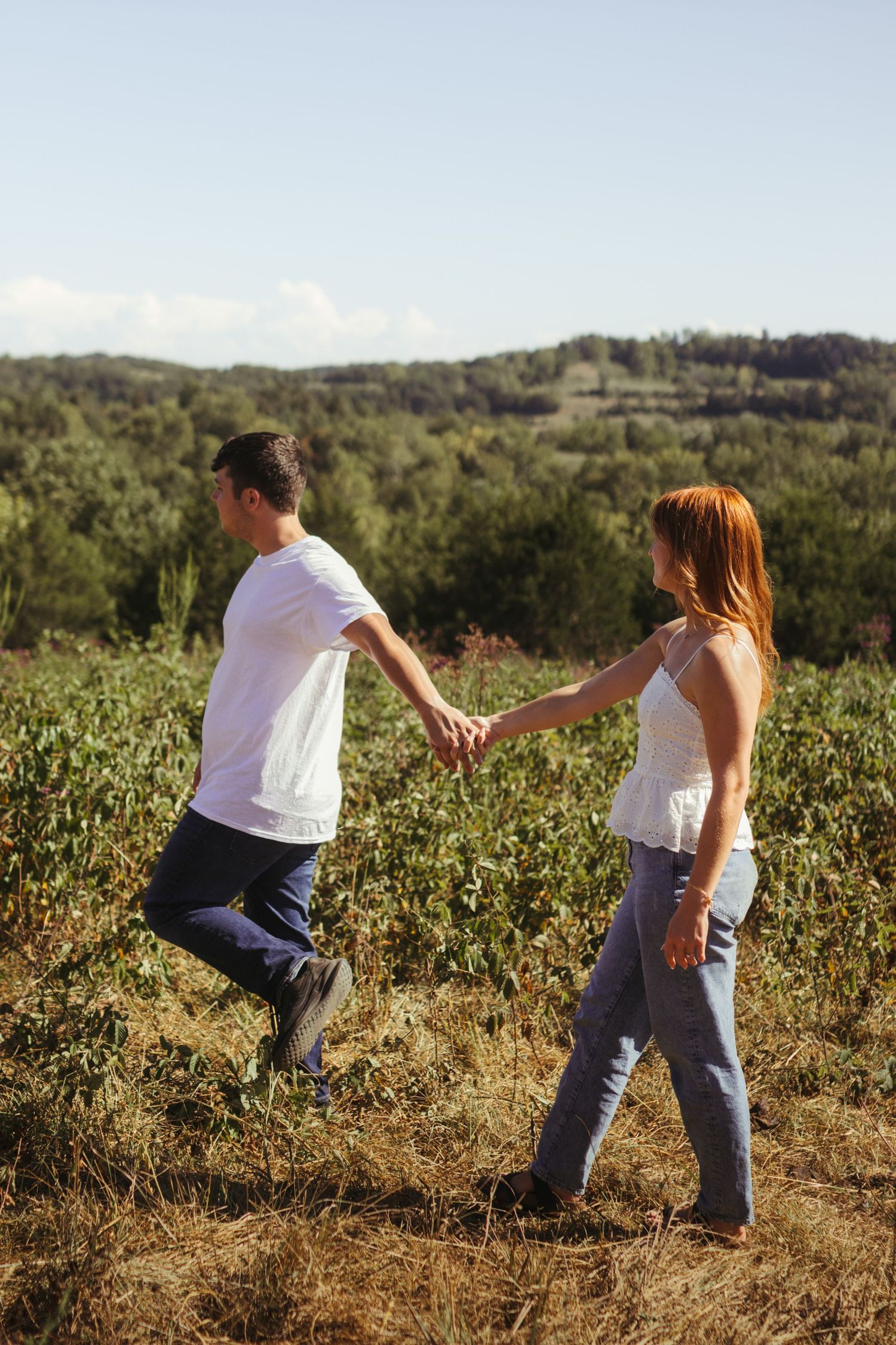 An engaged couple walking in a field with mountain view and beautiful blue sky. The couple is holding hands.