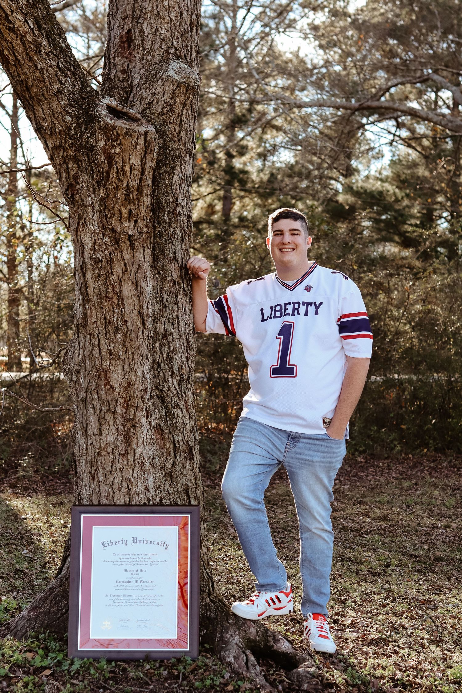 College senior portrait in the woods leaning against a tree with a Liberty university degree and jersey.