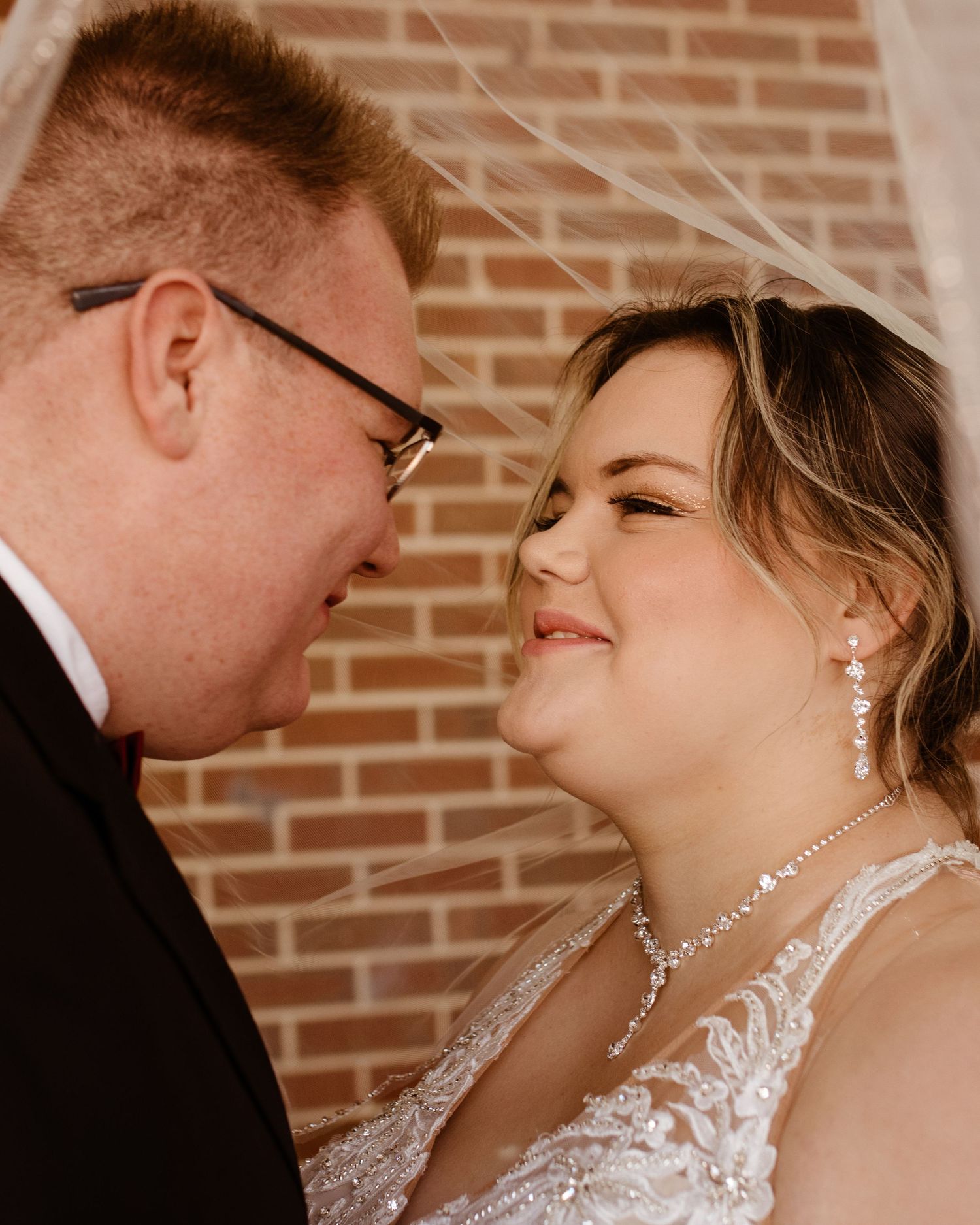 Couple on their wedding day under a veil and smiling at each other.