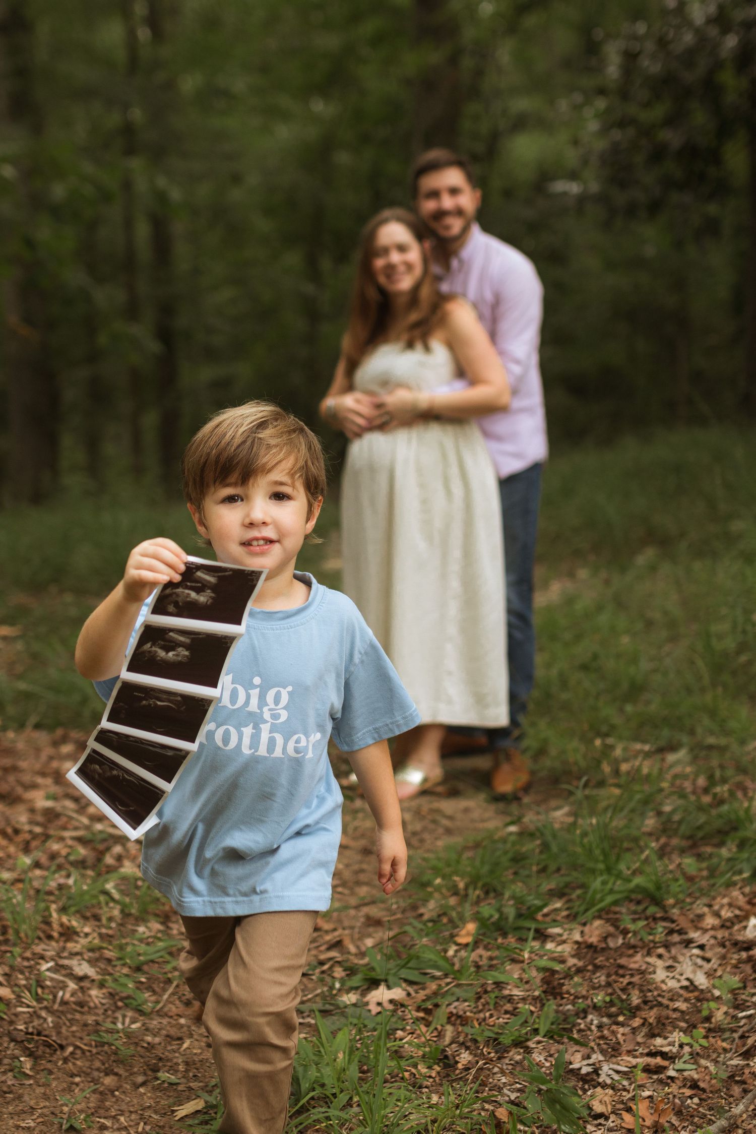 A young boy holding ultrasound photos and running towards the camera. The mom and dad are in the distance hugging and looking at the camera.