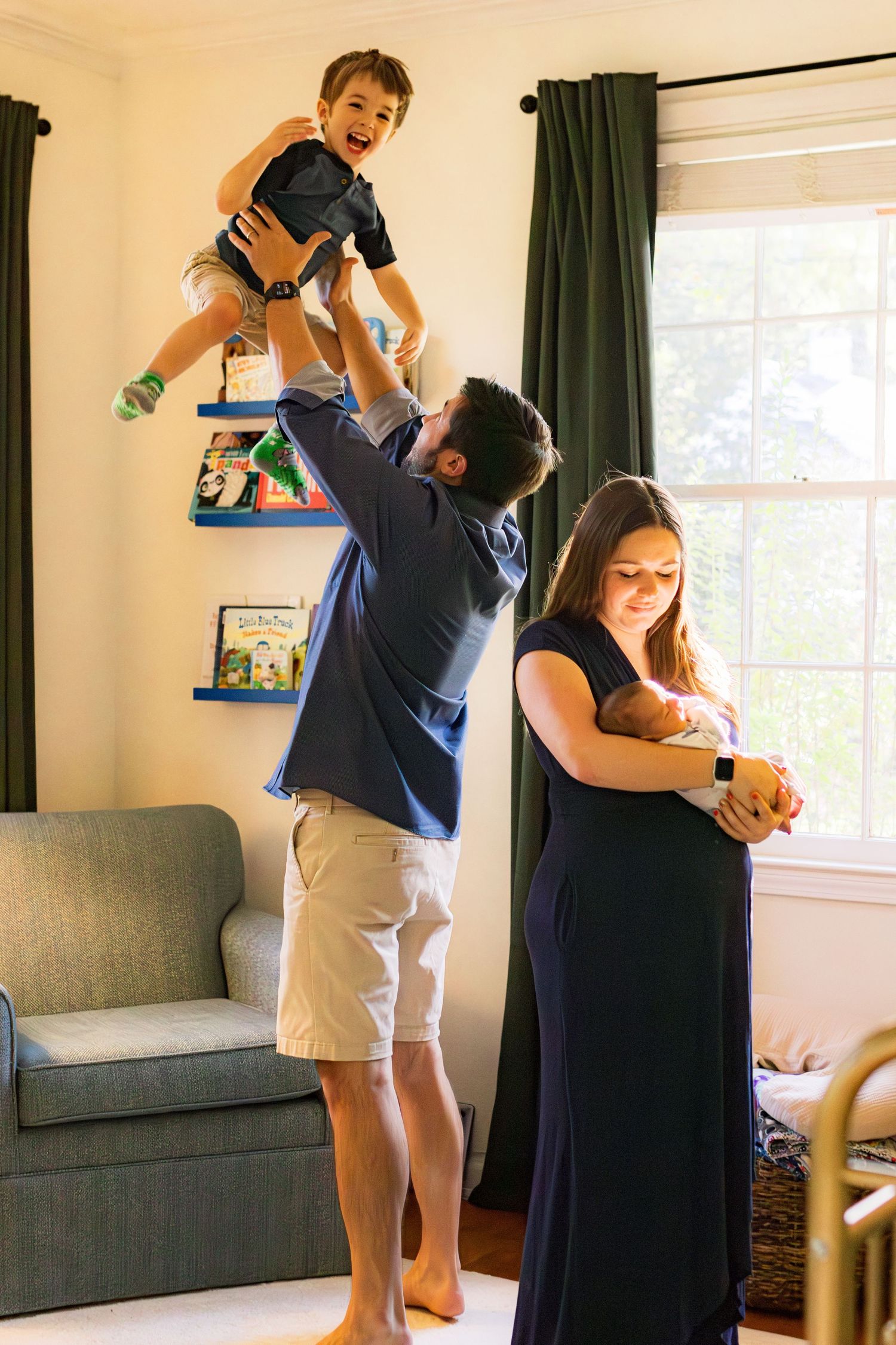 A family in a nursery in front of a window. A day is throwing a young boy in the air playfully and the mom is holding her newborn son.