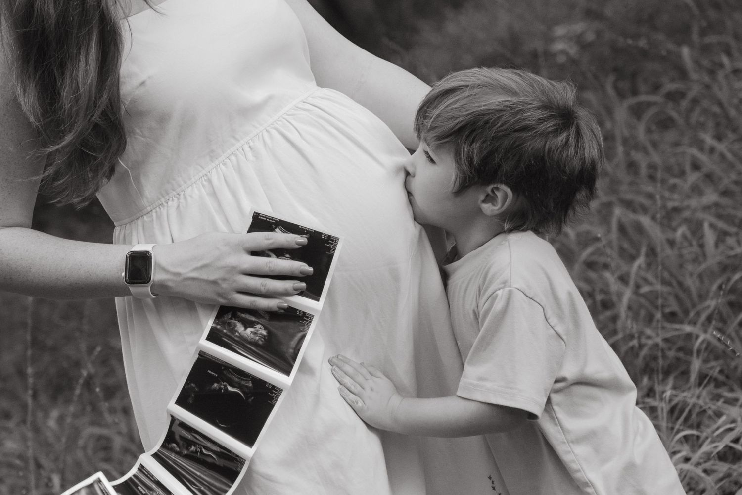 Pregnant woman holding ultrasound photos with her four year old son kissing her belly.