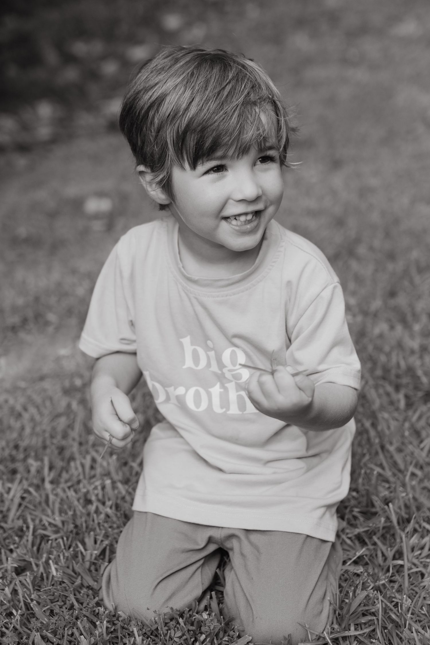Young boy sitting in the grass playing with grass with a shirt on that says 