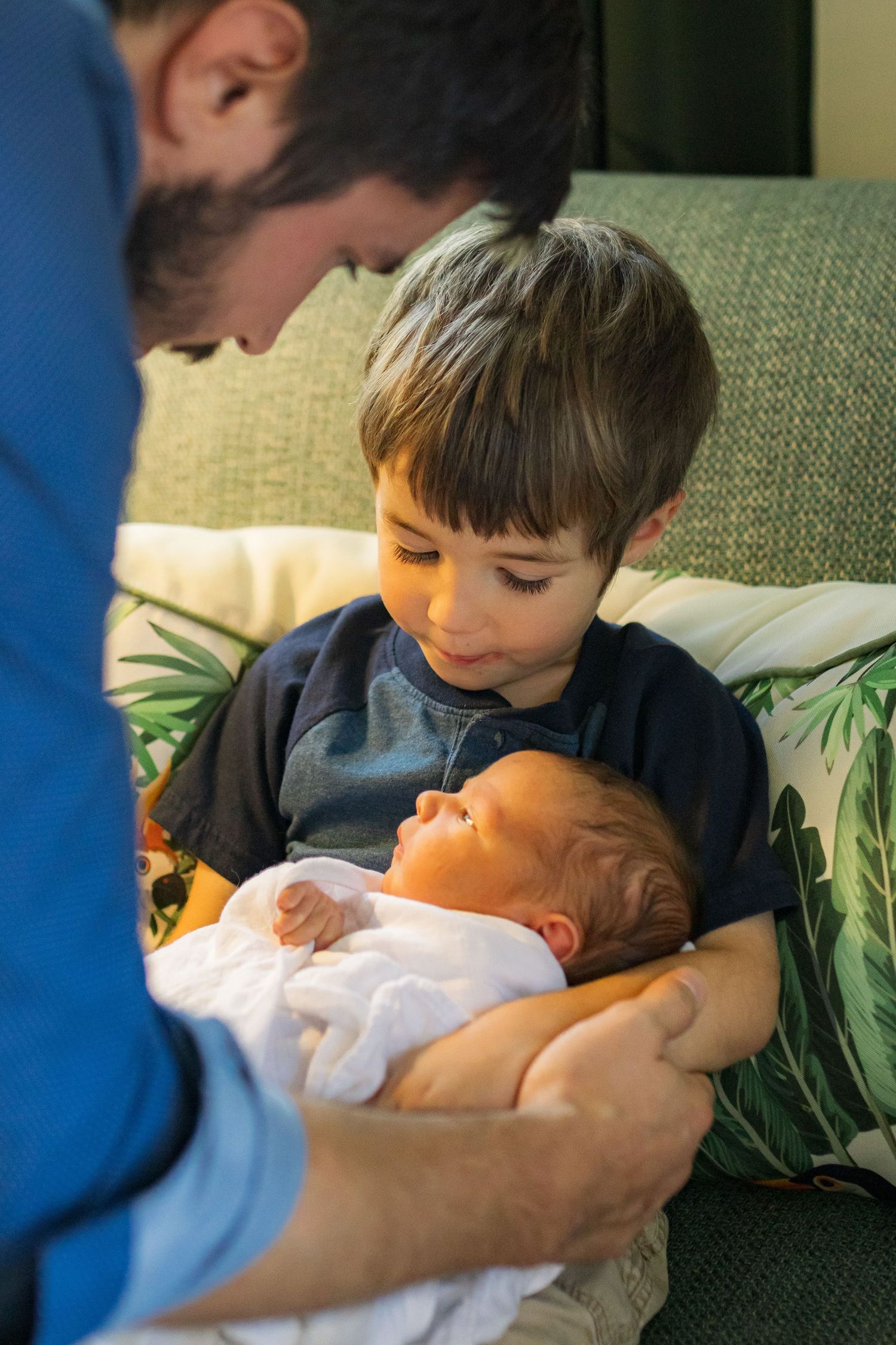 A dad handing his newborn son to his four year old son. The four year old is looking down at the newborn and smiling.