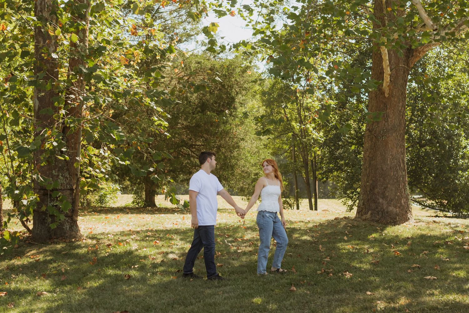 An engaged couple in a field with trees on either side walking and holding hands.