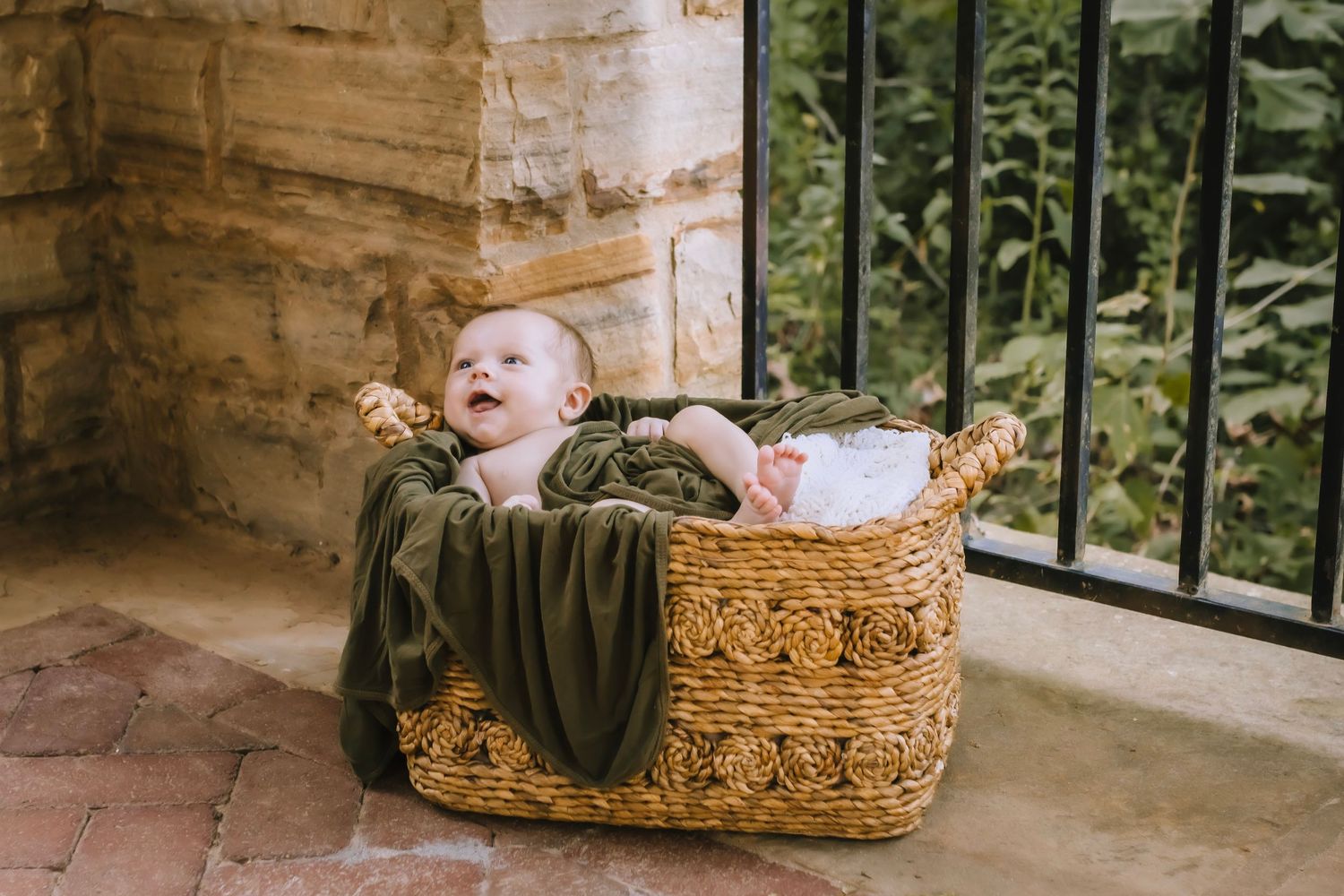 A newborn baby in a wicker basket with a green blanket. A stone wall and black guardrail in the background and brick floor. Smiling baby.