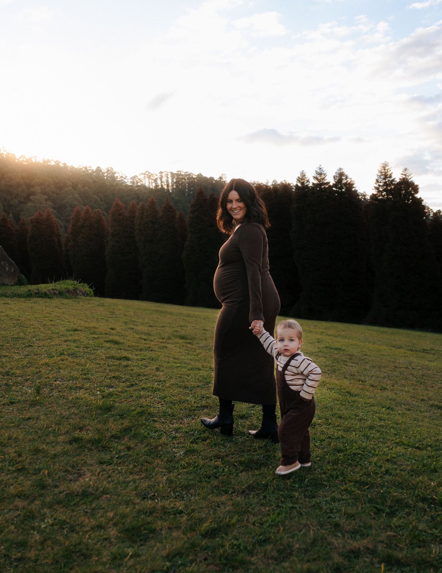 Pregnant woman in brown dress holding toddler's hand on green lawn at sunset.