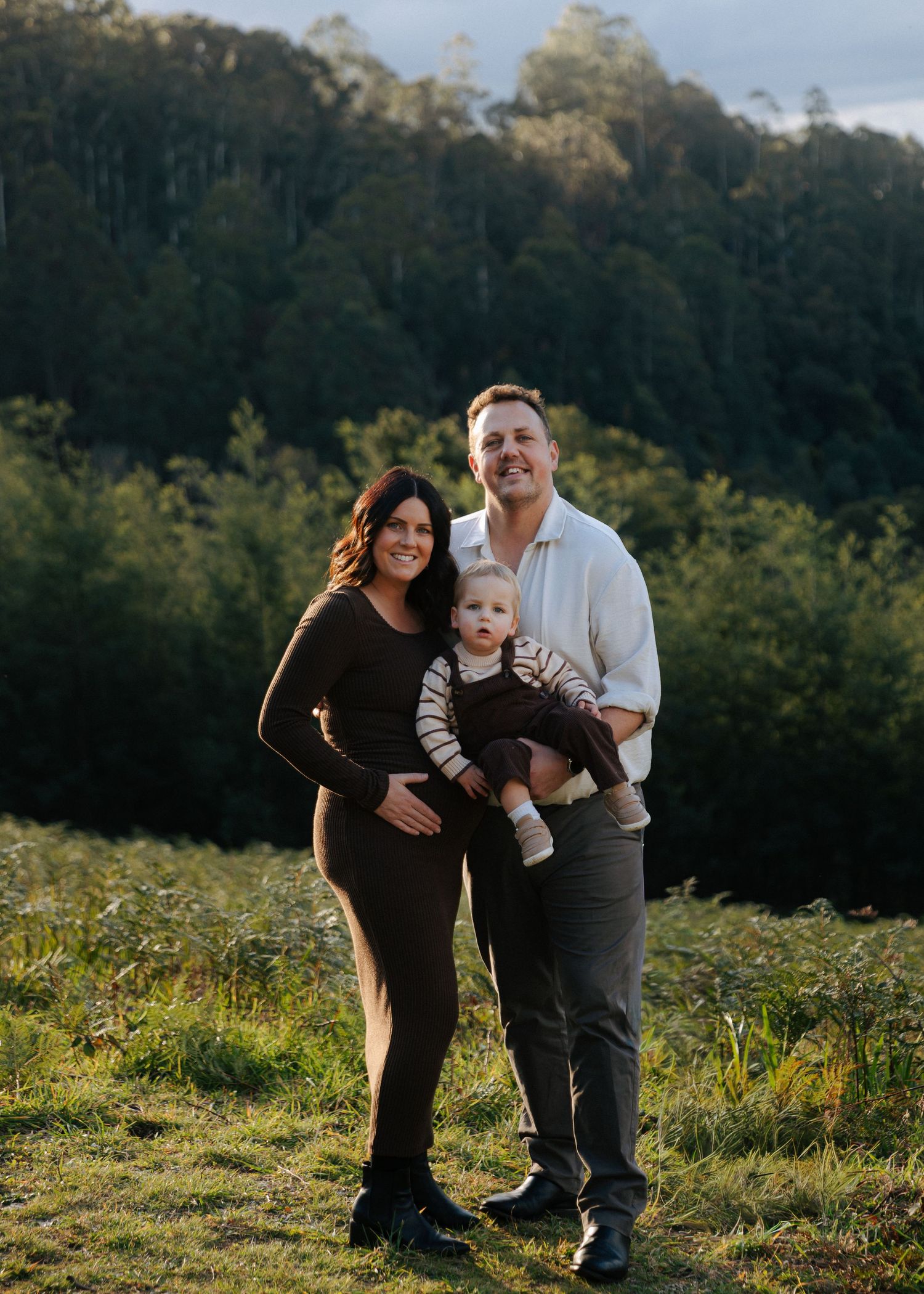 Smiling family portrait with pregnant mother, father holding toddler in golden light.