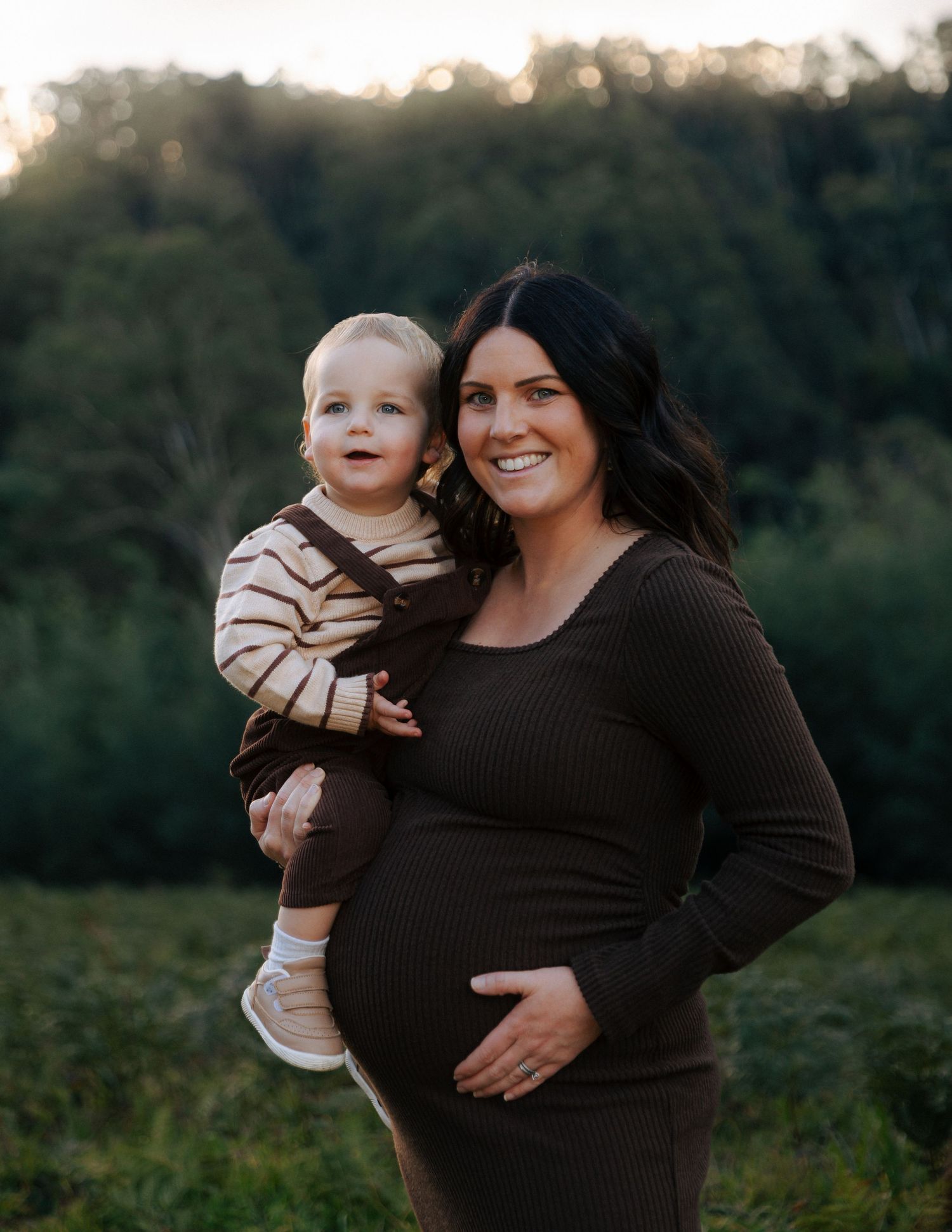 Smiling pregnant woman in brown ribbed dress holding a happy toddler in a field at sunset.