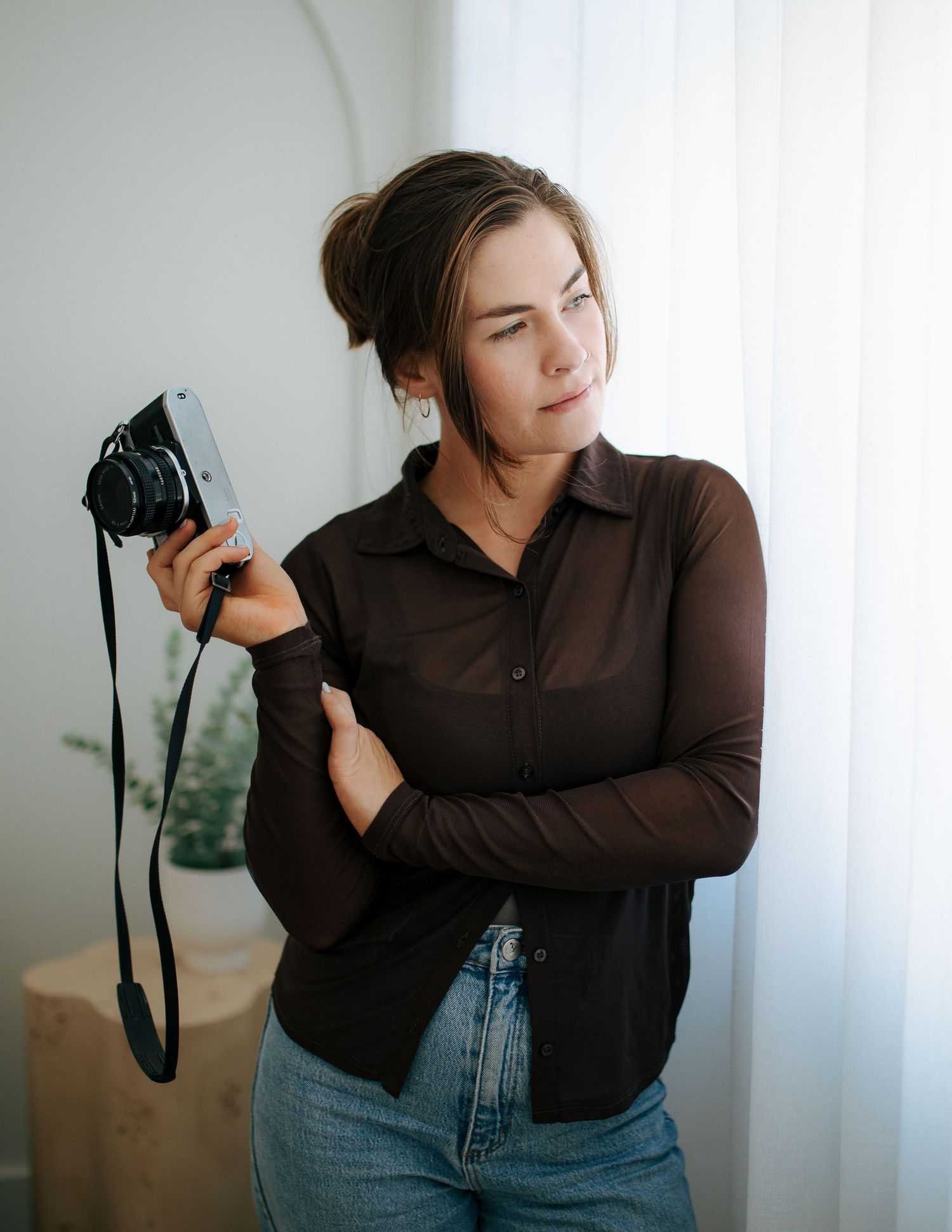 Young woman in brown shirt holding vintage film camera near bright window.