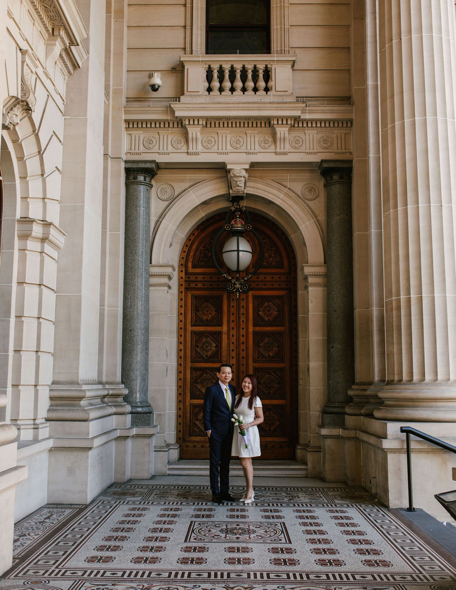 Couple standing before ornate arched wooden door of classical heritage building with mosaic floor.