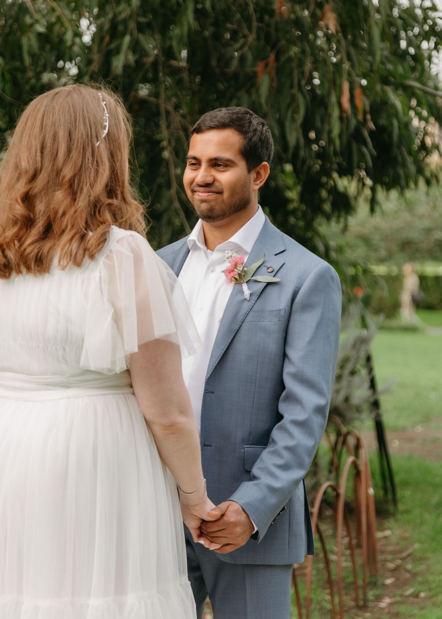 Groom in blue suit smiles at bride during outdoor wedding ceremony.