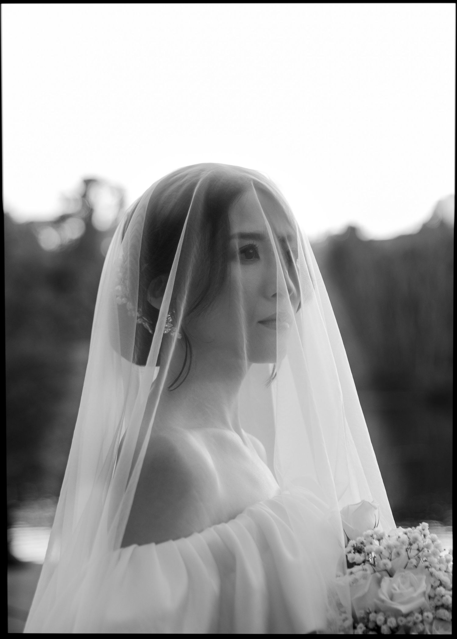Black and white portrait of a bride draped in a sheer veil, holding a bouquet in soft natural light.