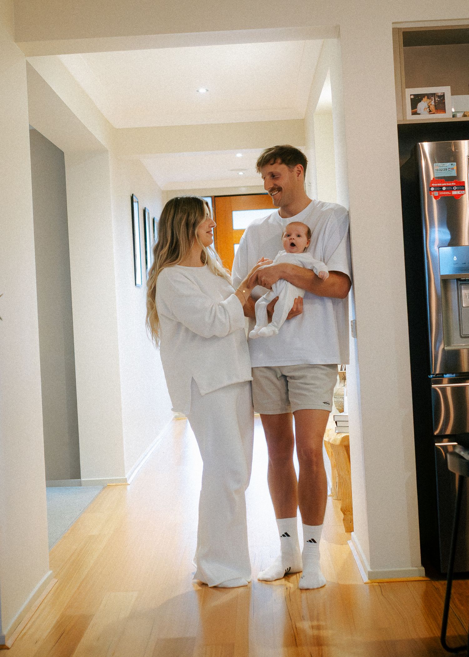 Happy couple in white outfits standing in bright hallway holding their newborn baby.