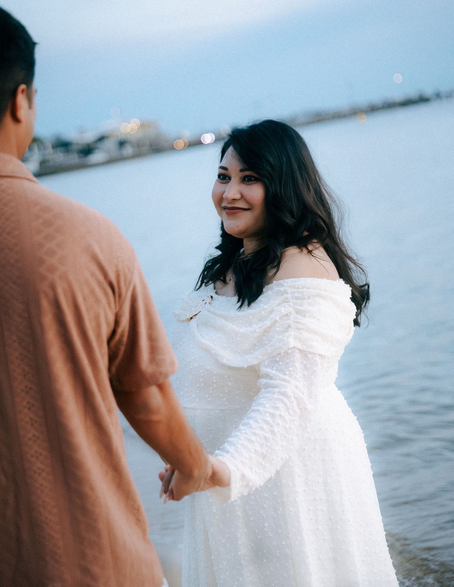 Pregnant woman in white off-shoulder dress smiling while holding hands with partner by water.