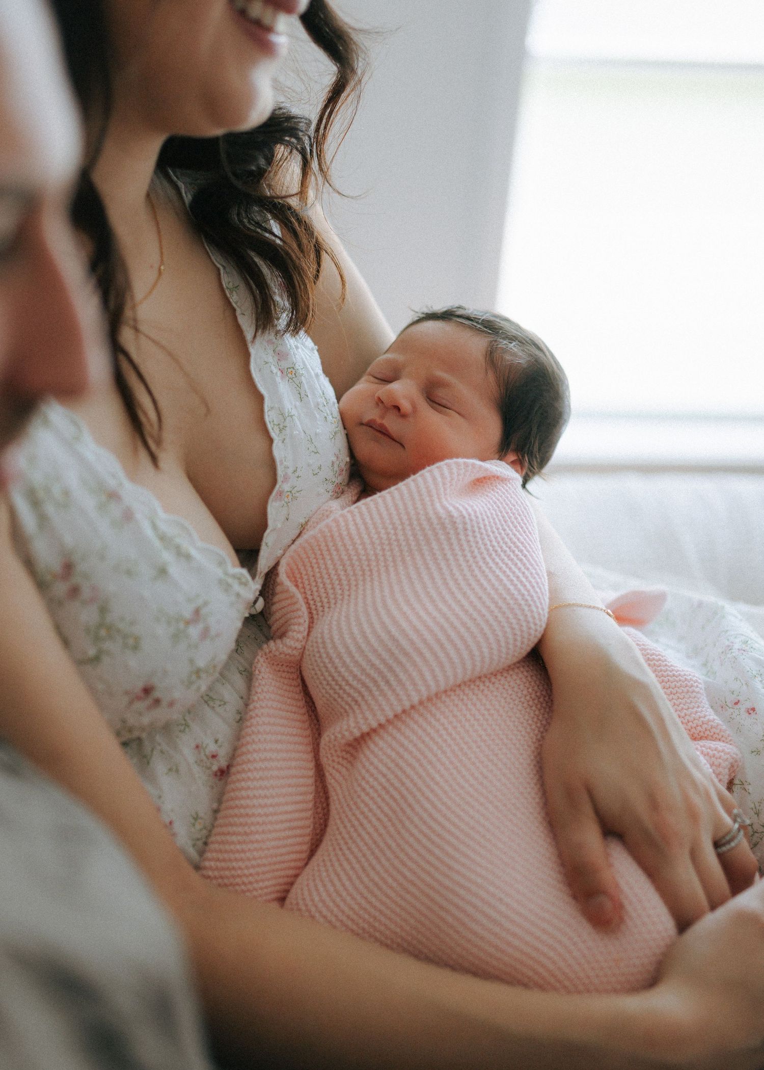 Mother in floral dress tenderly holding sleeping newborn baby wrapped in pink blanket.