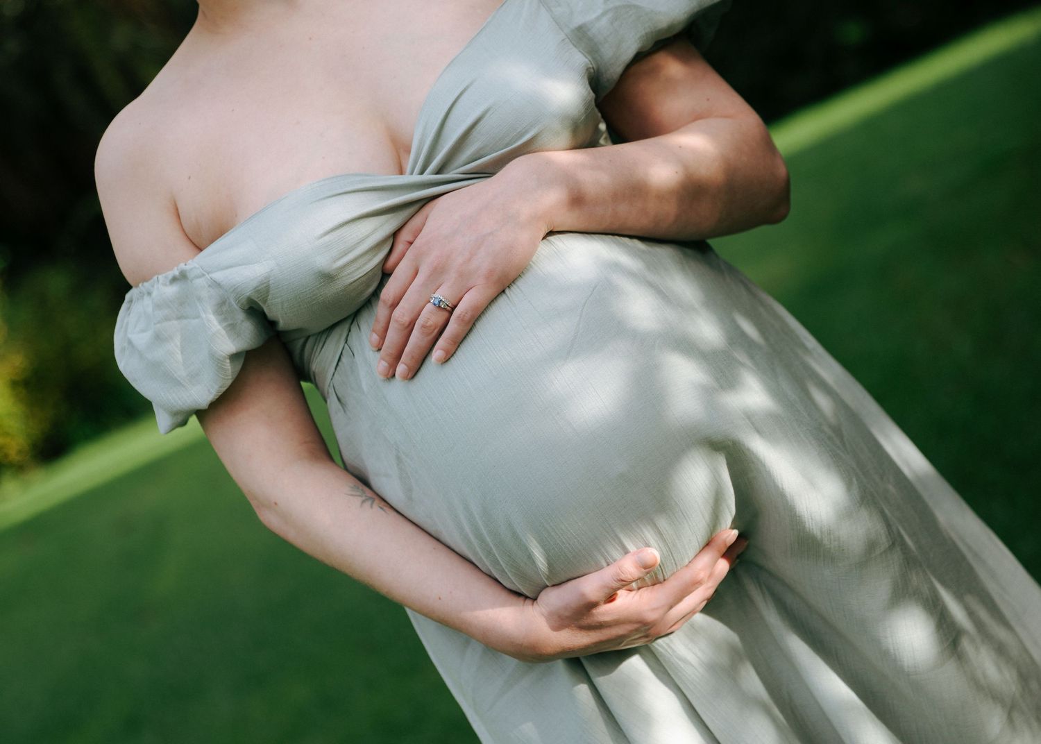 Close-up of pregnant woman in sage green dress with partner's hands gently holding baby bump outdoors.