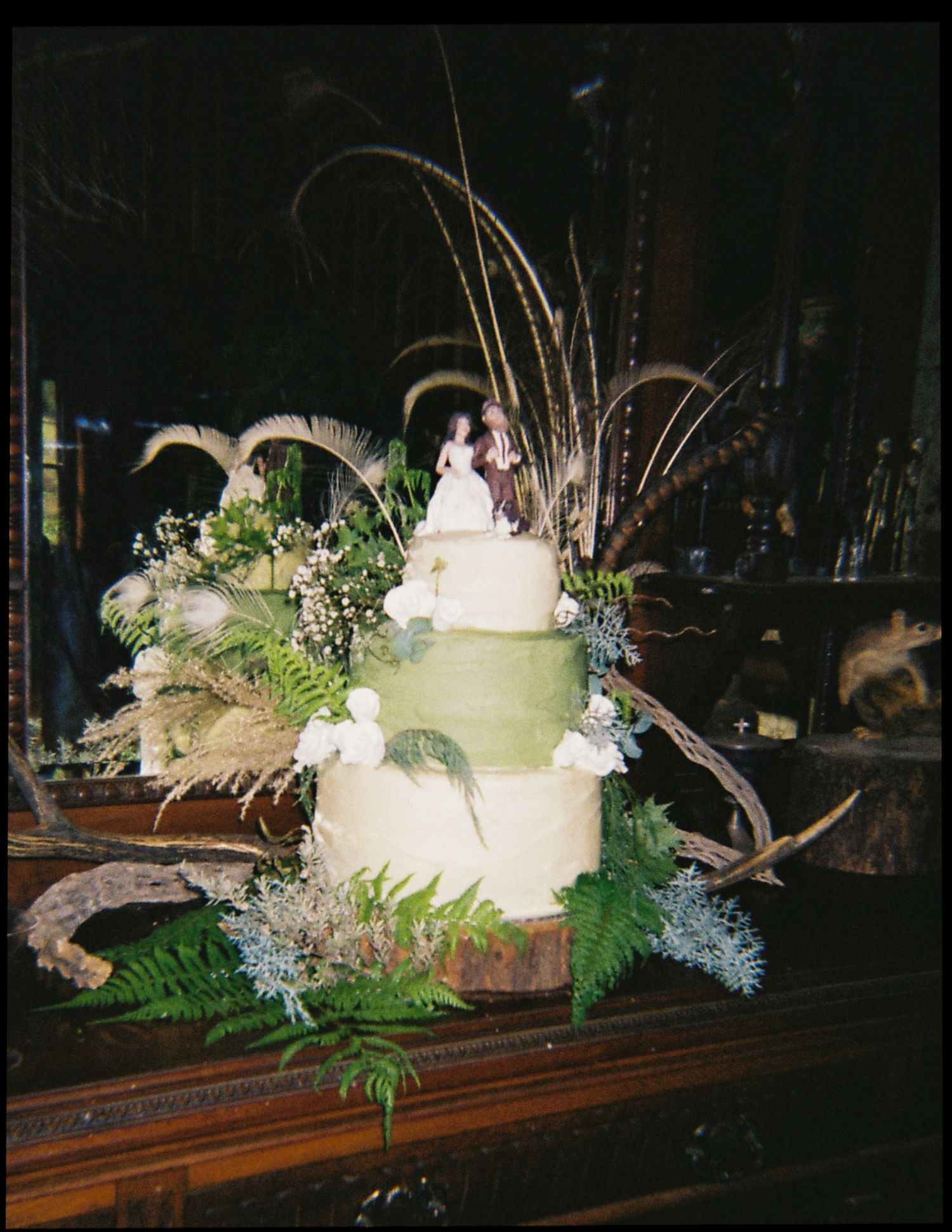 Elegant two-tier white wedding cake decorated with ferns, feathers, and a bride and groom cake topper.