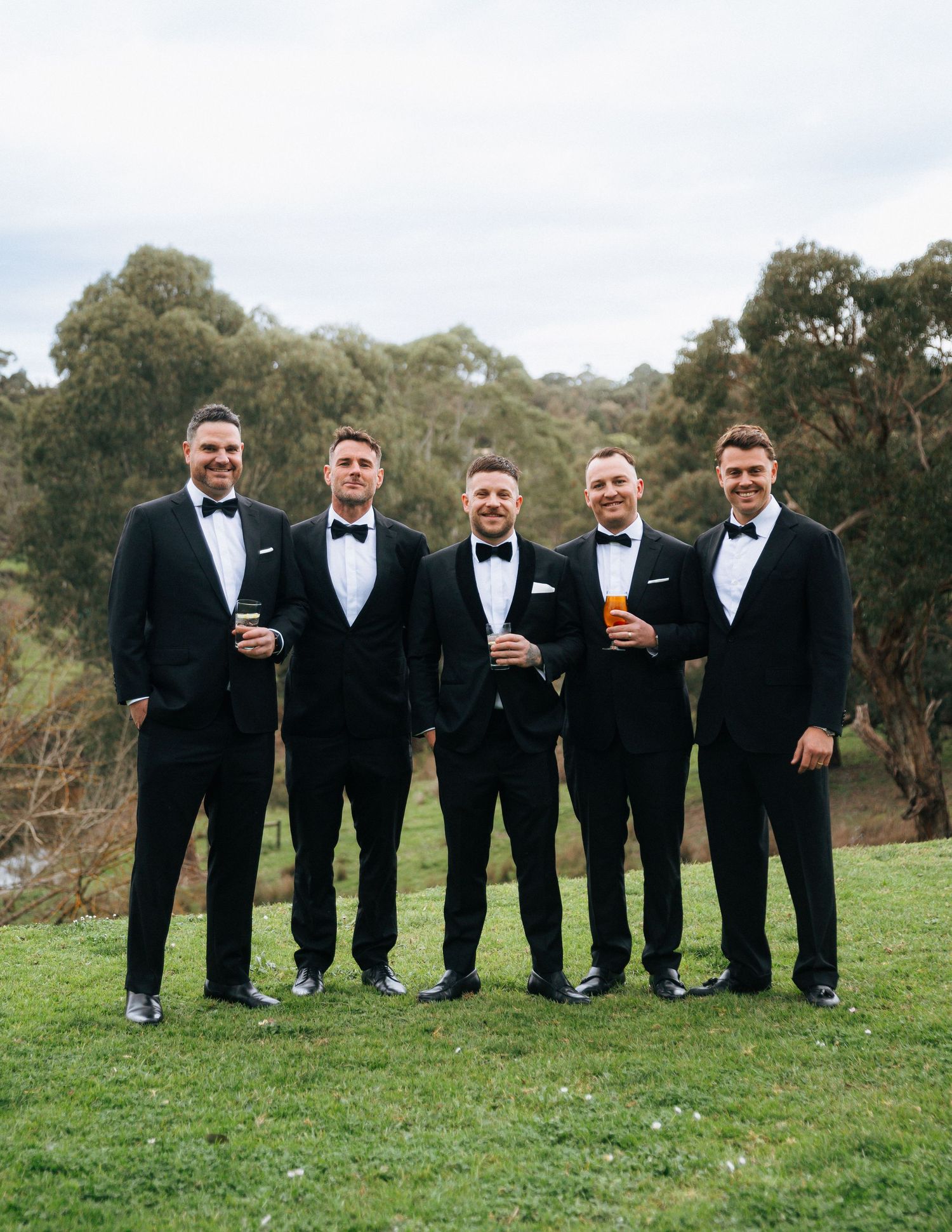Five groomsmen in black tuxedos and bow ties holding drinks while posing outdoors on green grass with trees behind.