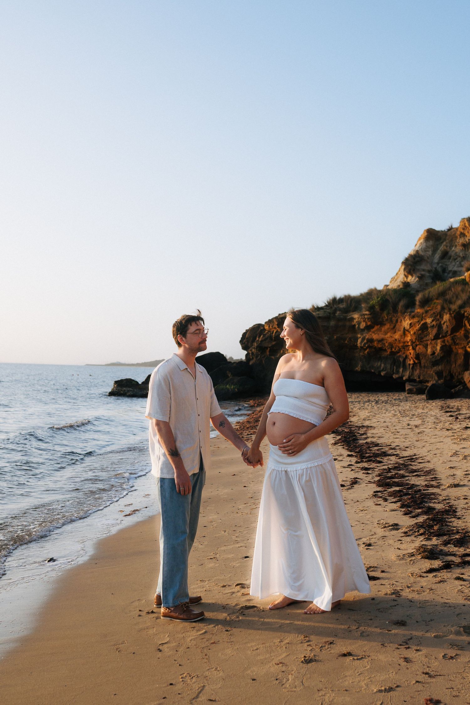 A couple holds hands on a sandy beach at sunset, the woman in a white outfit cradling her pregnant belly.
