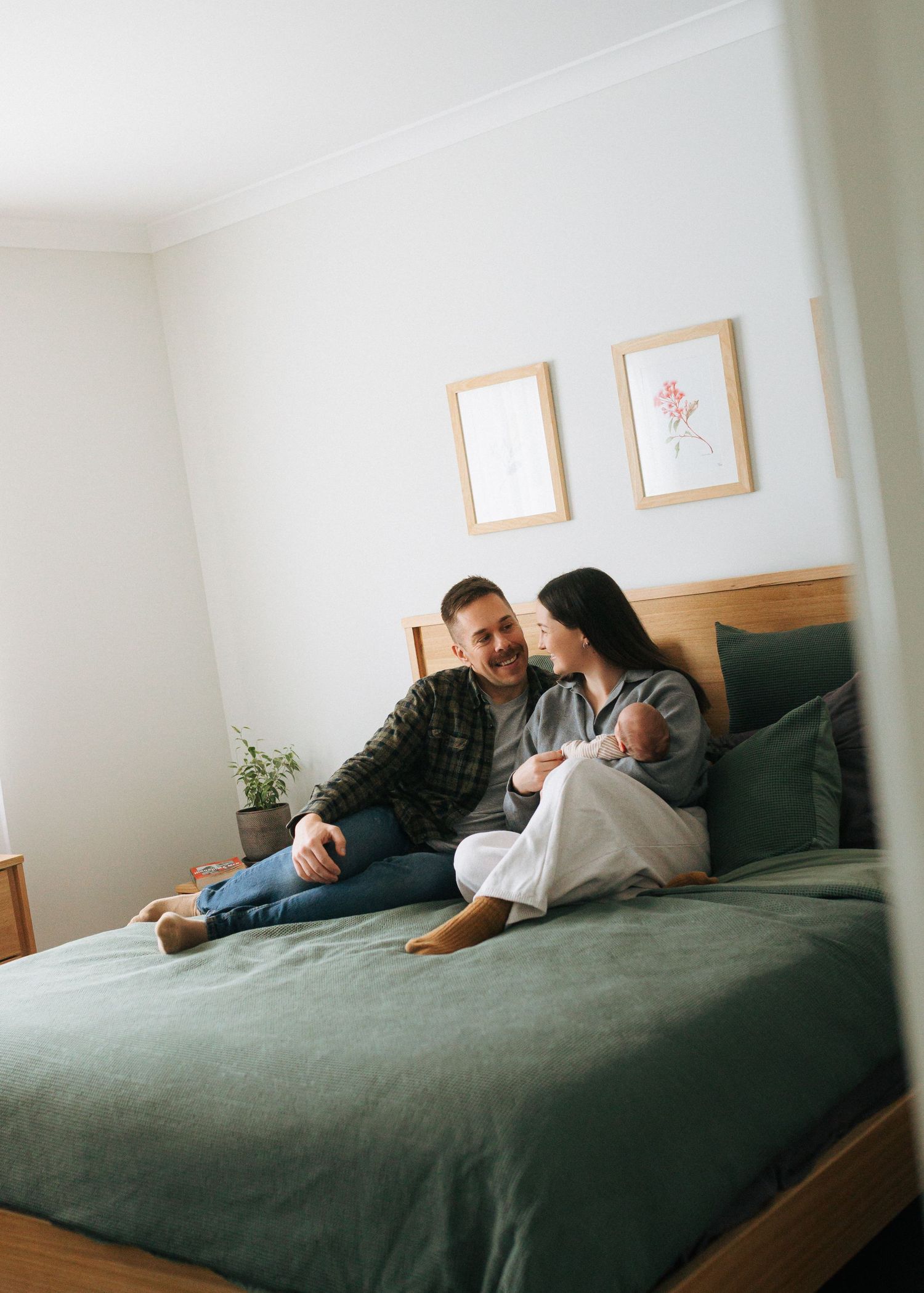 A couple sits on a green bed laughing together while holding their newborn baby at home.