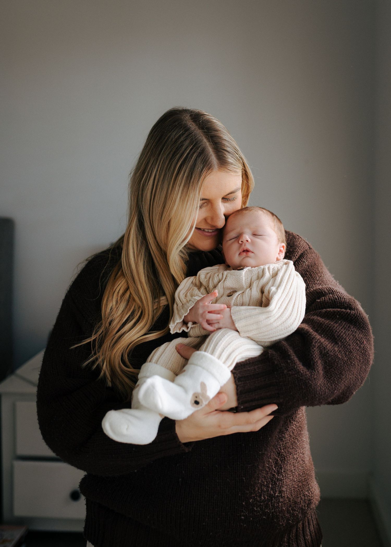Mother in brown sweater holding sleeping newborn baby dressed in cream knit outfit and white booties.