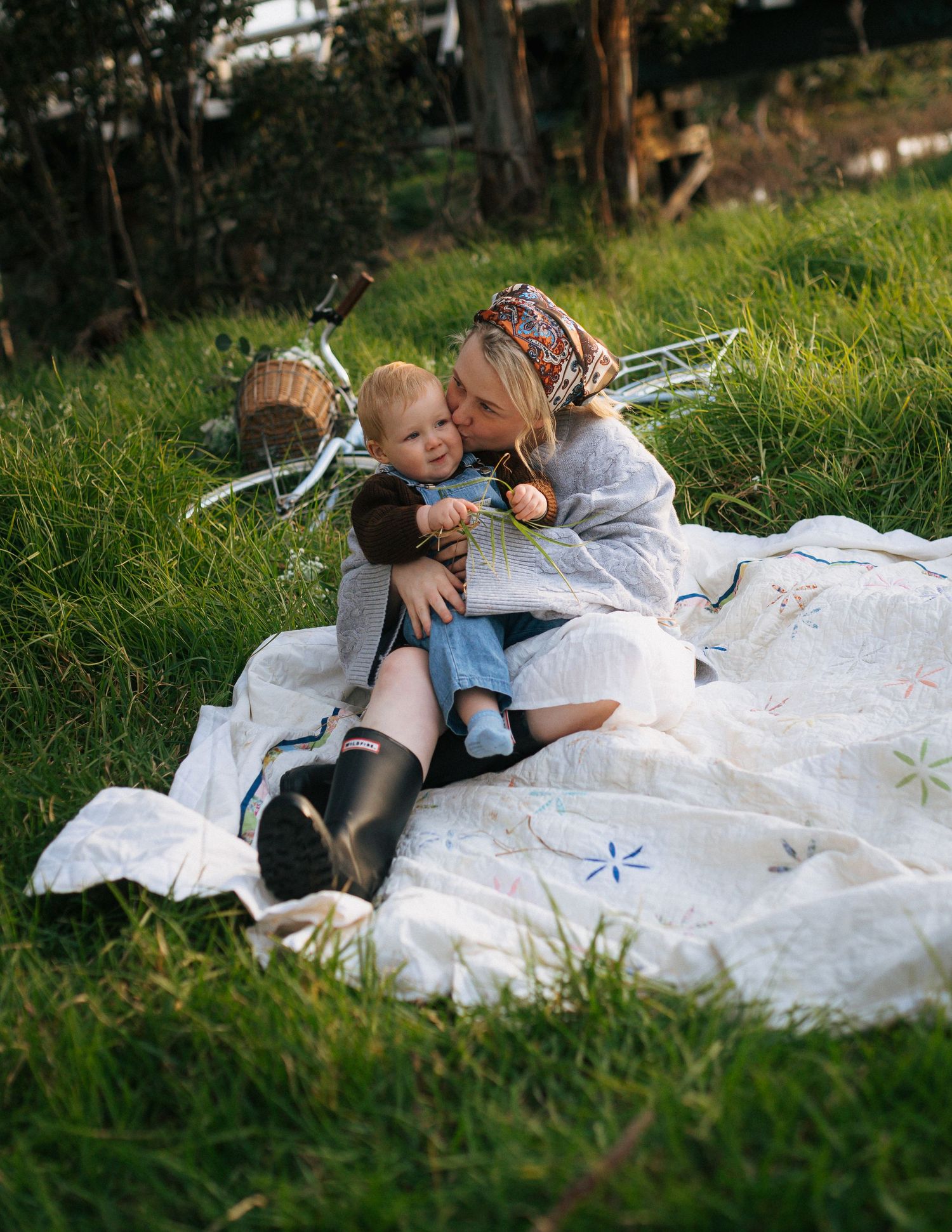 Mother kissing her smiling toddler while sitting on a white quilt in a green meadow.