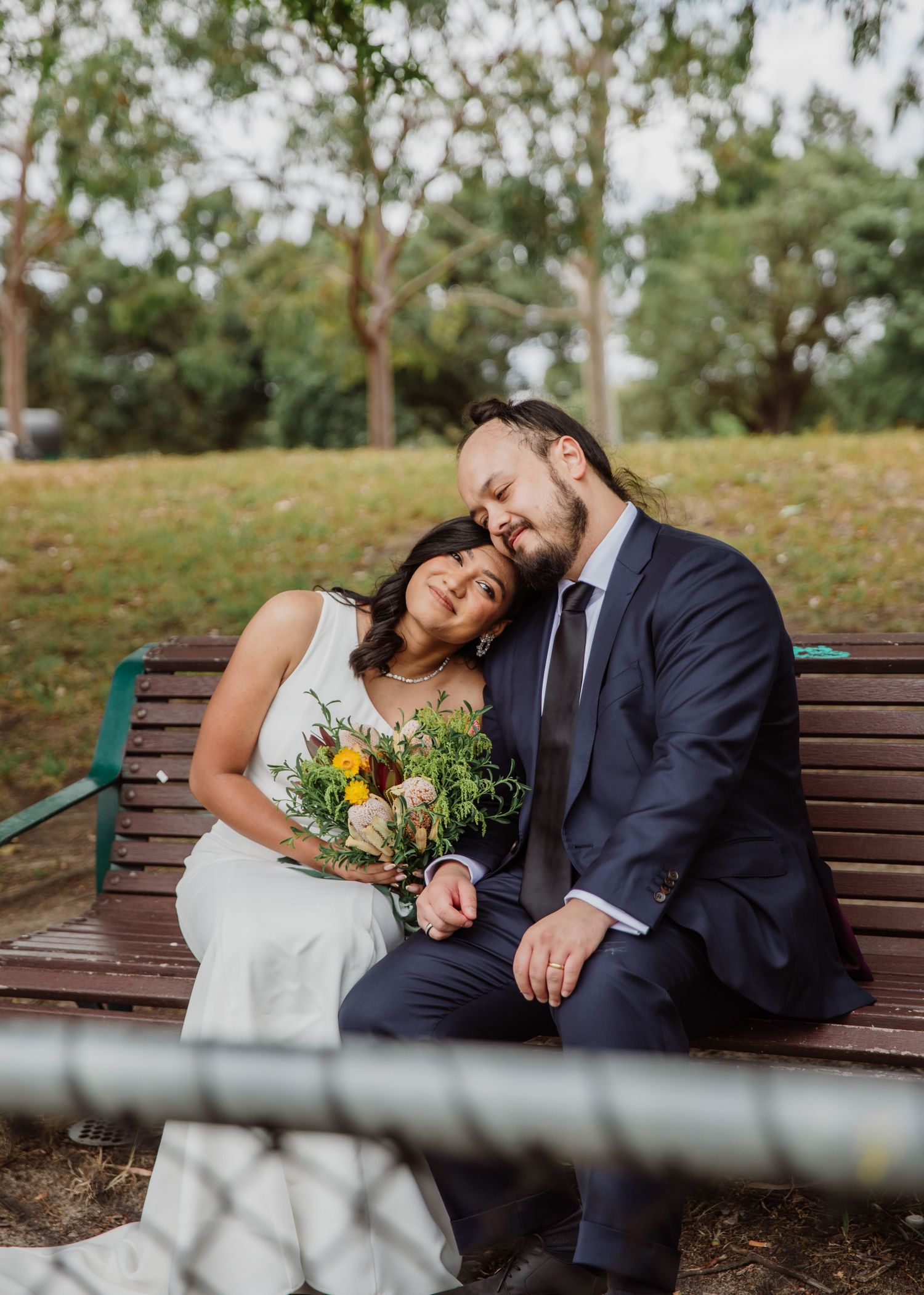 Bride and groom sitting closely on a park bench, bride holding a yellow bouquet.