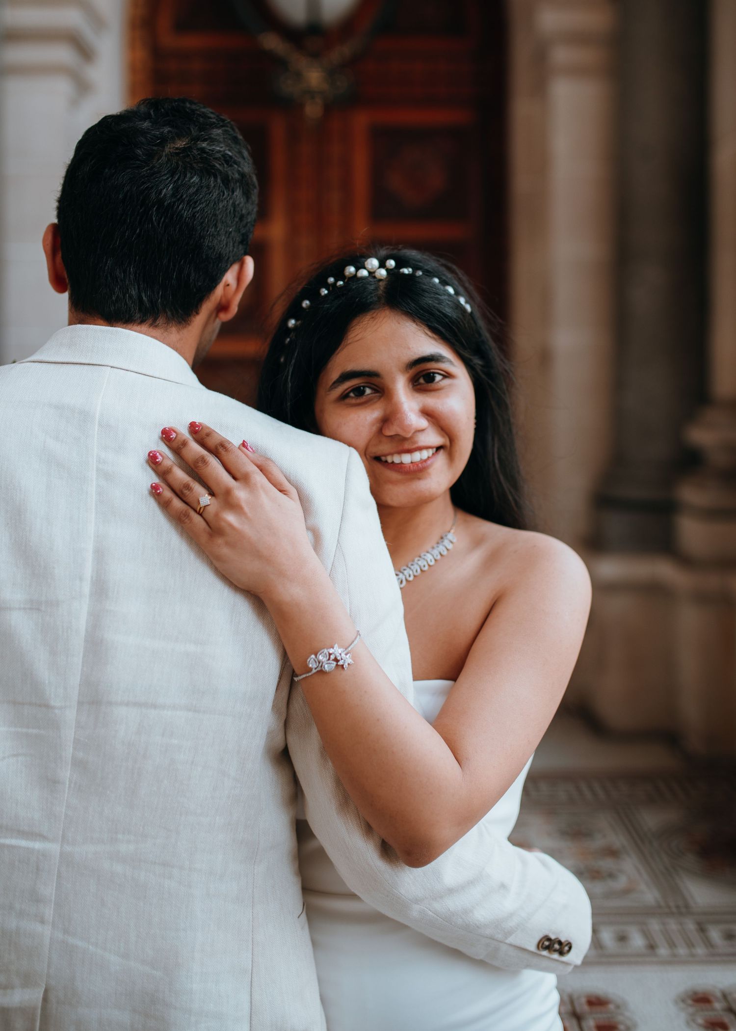 Smiling bride in white strapless gown with pearl headpiece and diamond necklace embraces groom in white suit.