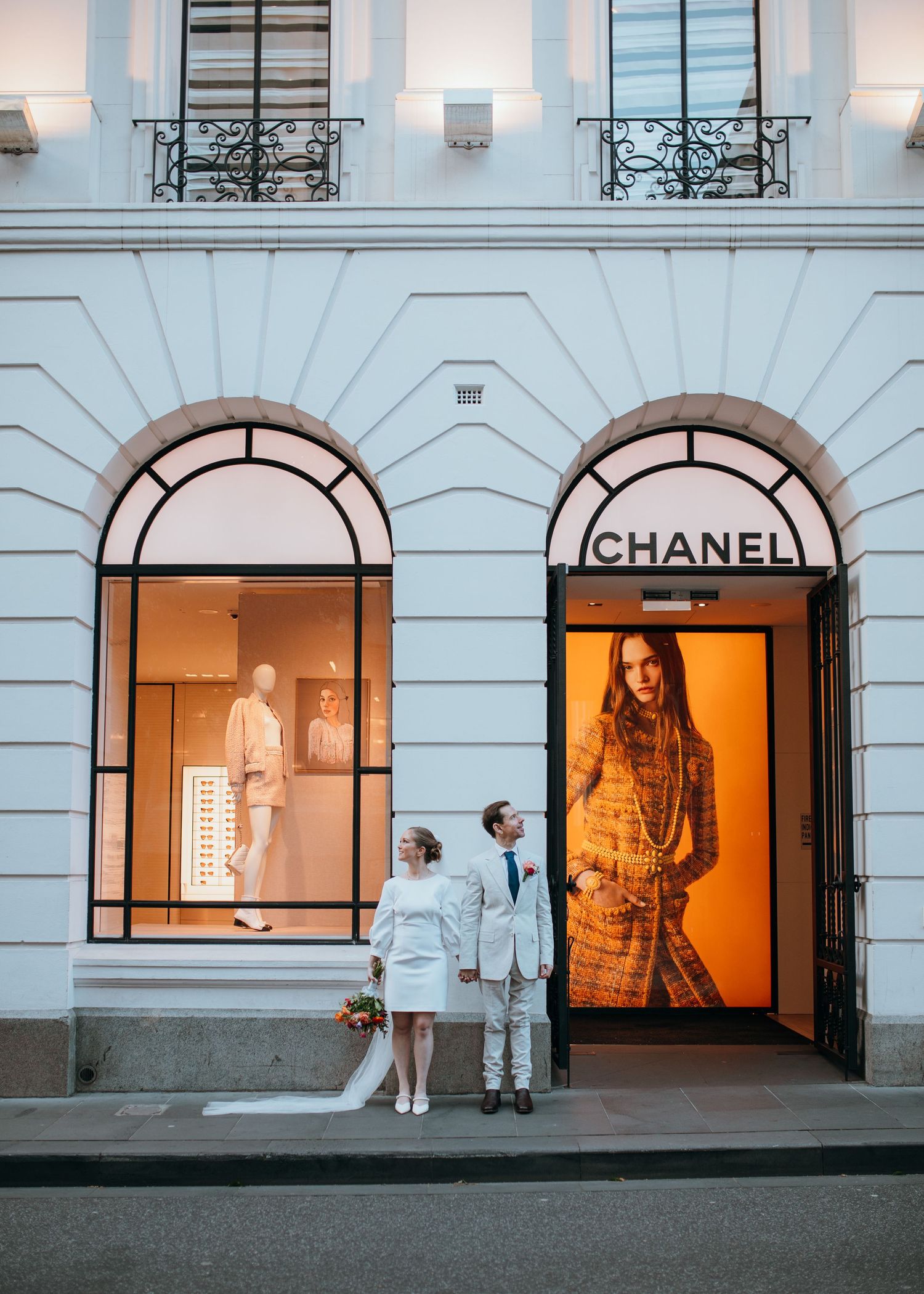 Bride and groom posing outside elegant Chanel boutique entrance with glowing orange display window.