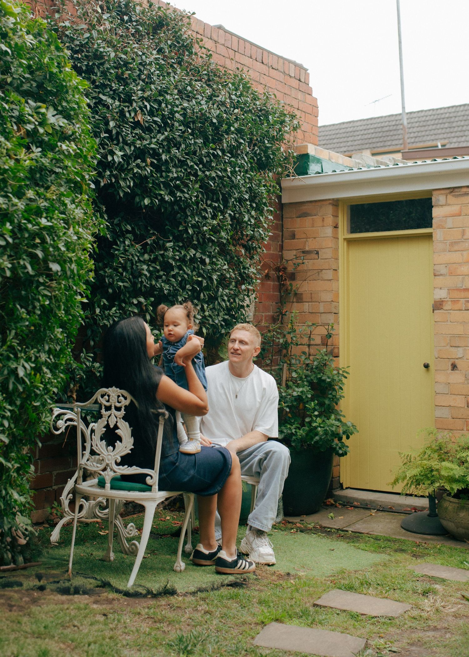 Family of three relaxing in a lush backyard garden beside a brick home with a yellow door.