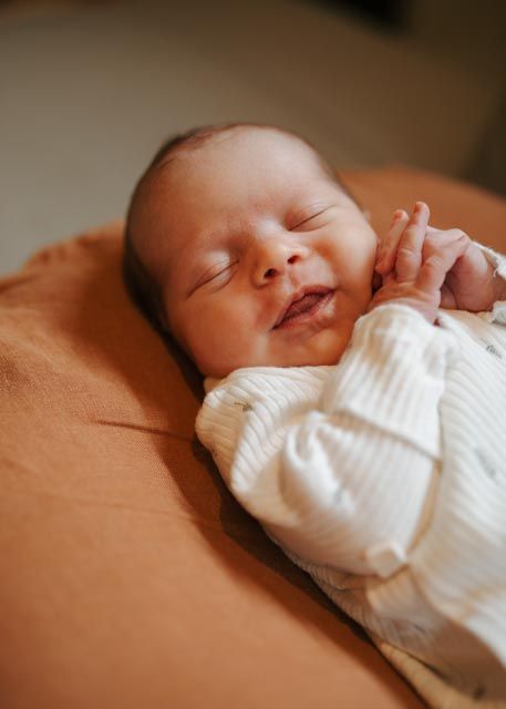 Peaceful newborn baby smiling softly while sleeping on rust-colored pillow in white ribbed onesie.